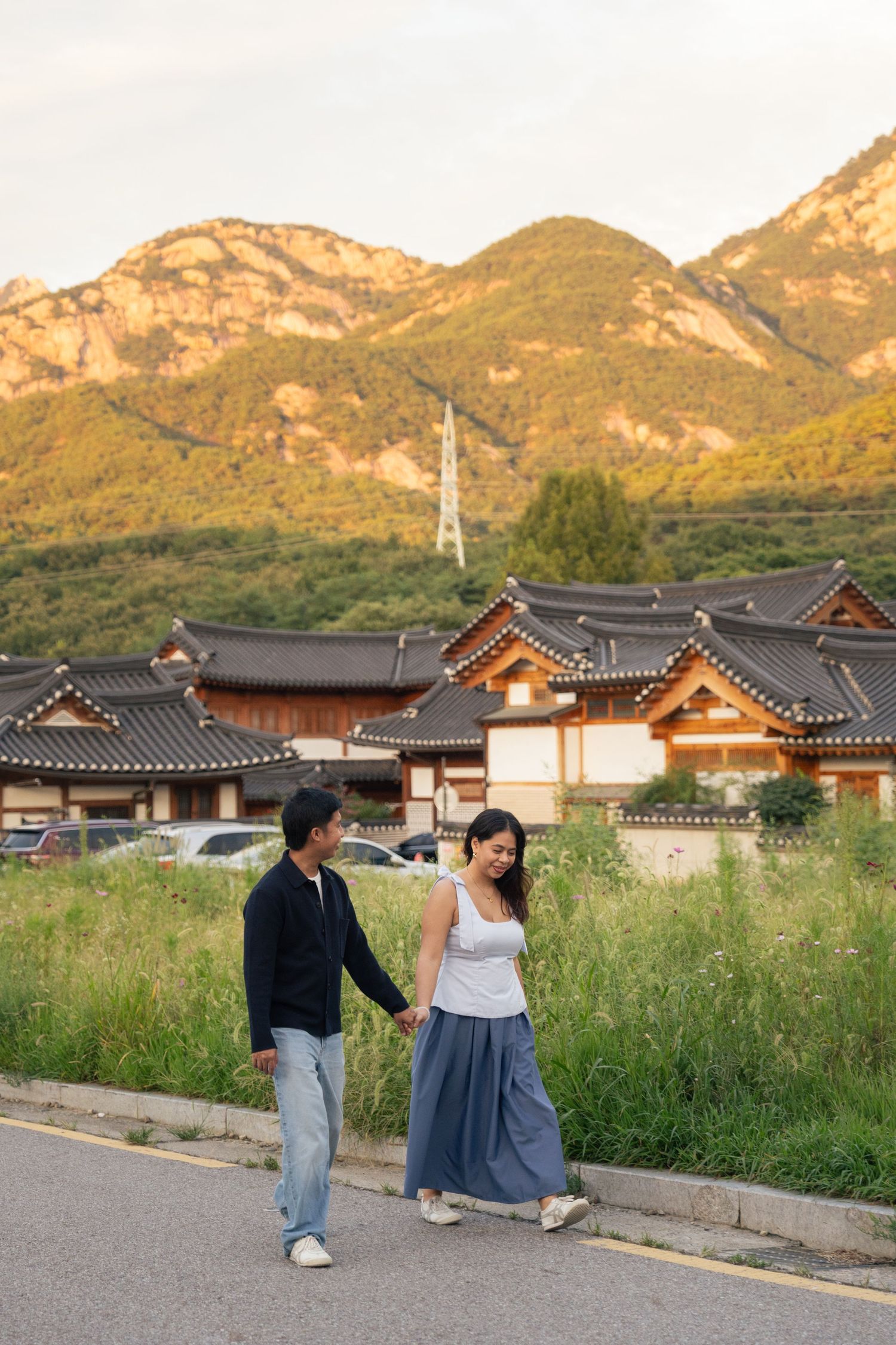 A couple walks along a road with traditional Korean hanok buildings and mountains in the background at sunset.