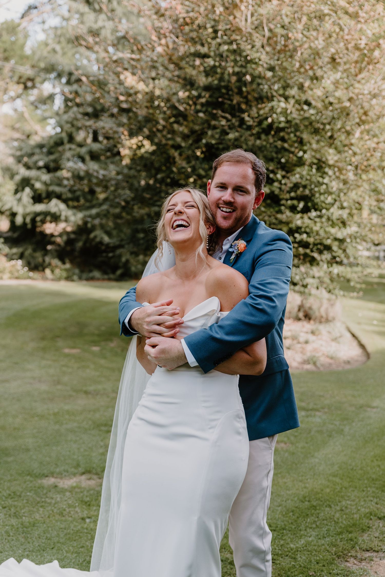 A couple shares joyful laughter during their wedding photo session on a lush green lawn surrounded by trees. christchurch wedding photograph