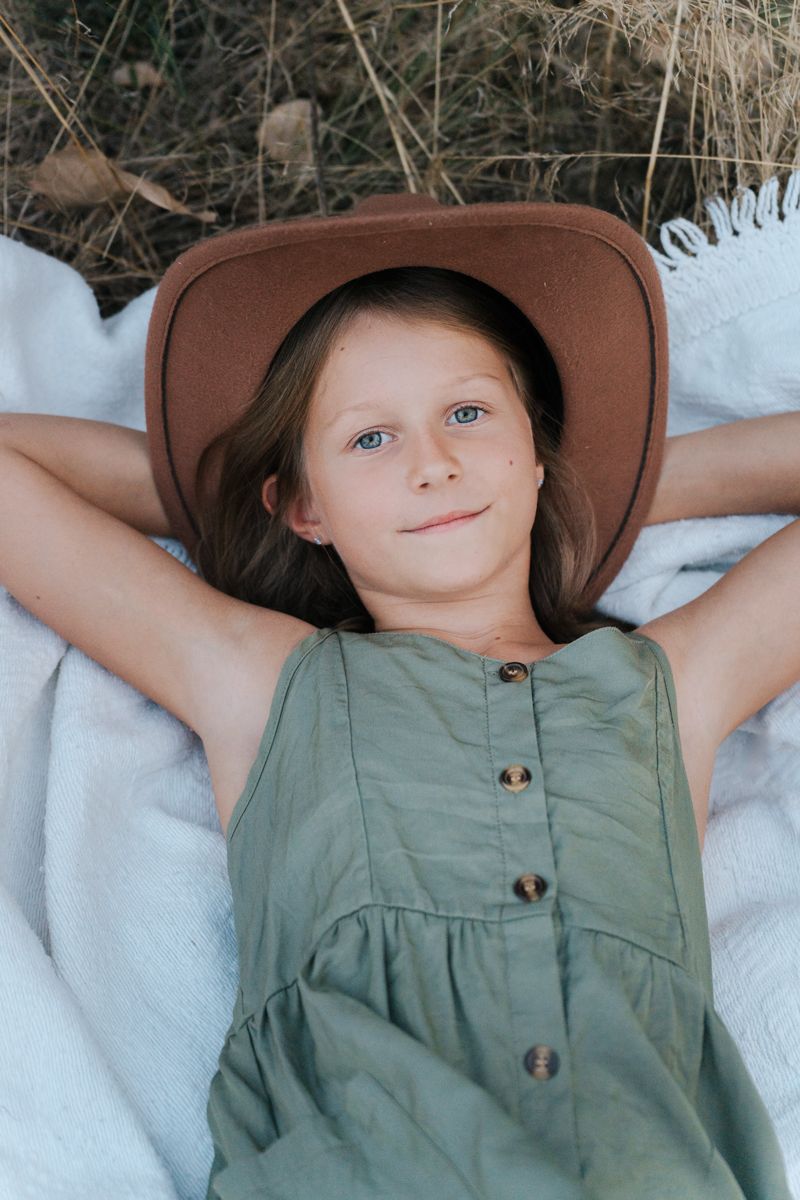 A child wearing a brown hat and sage green dress lies peacefully on white linens in three similar poses during a family photoshoot in Sydney