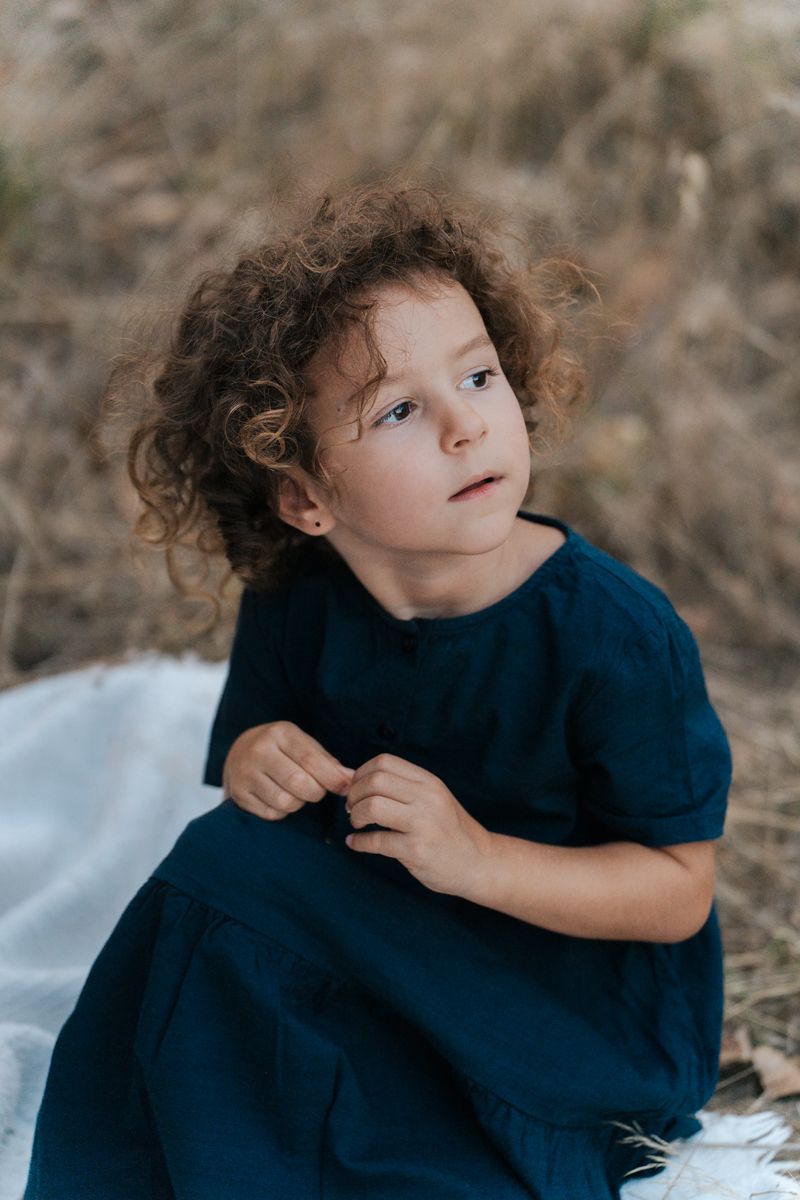 Child wearing navy blue dress sits on a white blanket outdoors while gazing upward with curly brown hair during family photoshoot in Sydney