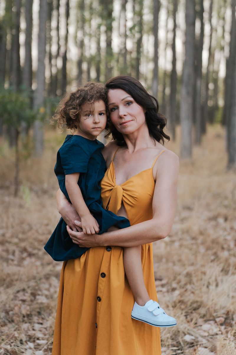 Mom in a yellow dress embraces their child in a navy outfit while standing among tall trees in western sydney parklands