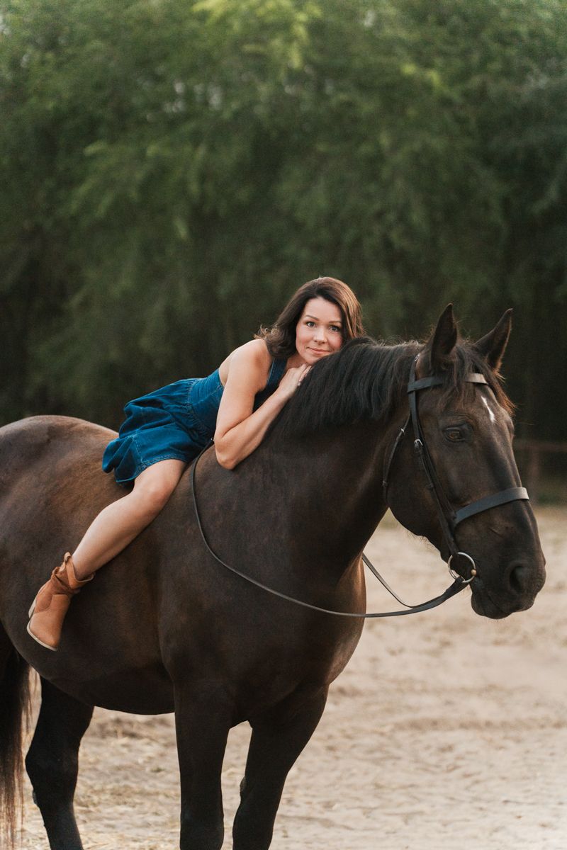 Woman in blue clothing lying on a dark horse's back in an outdoor setting with trees in the background during a horse photoshoot in Sydney
