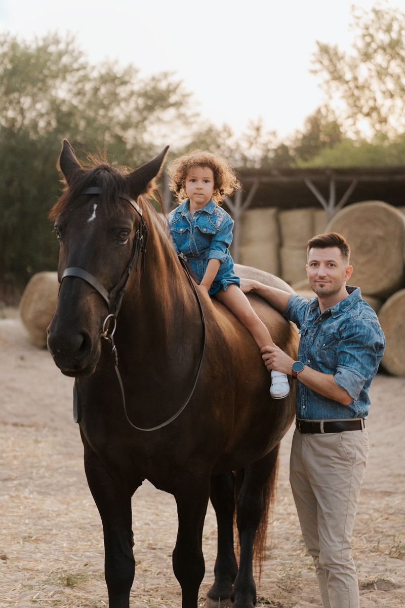 little girl sitting on a dark horse while her dad holds her leg and smiles towards the camera during a family photoshoot with horse in Hills