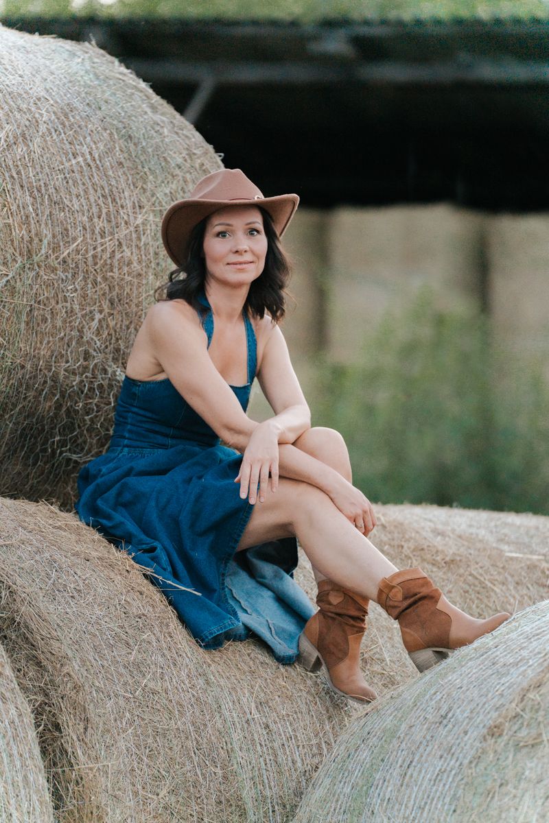 Woman wearing a denim dress and brown cowgirl boots sits on a large hay bales in a rustic outdoor setting  Western Sydney photoshoot