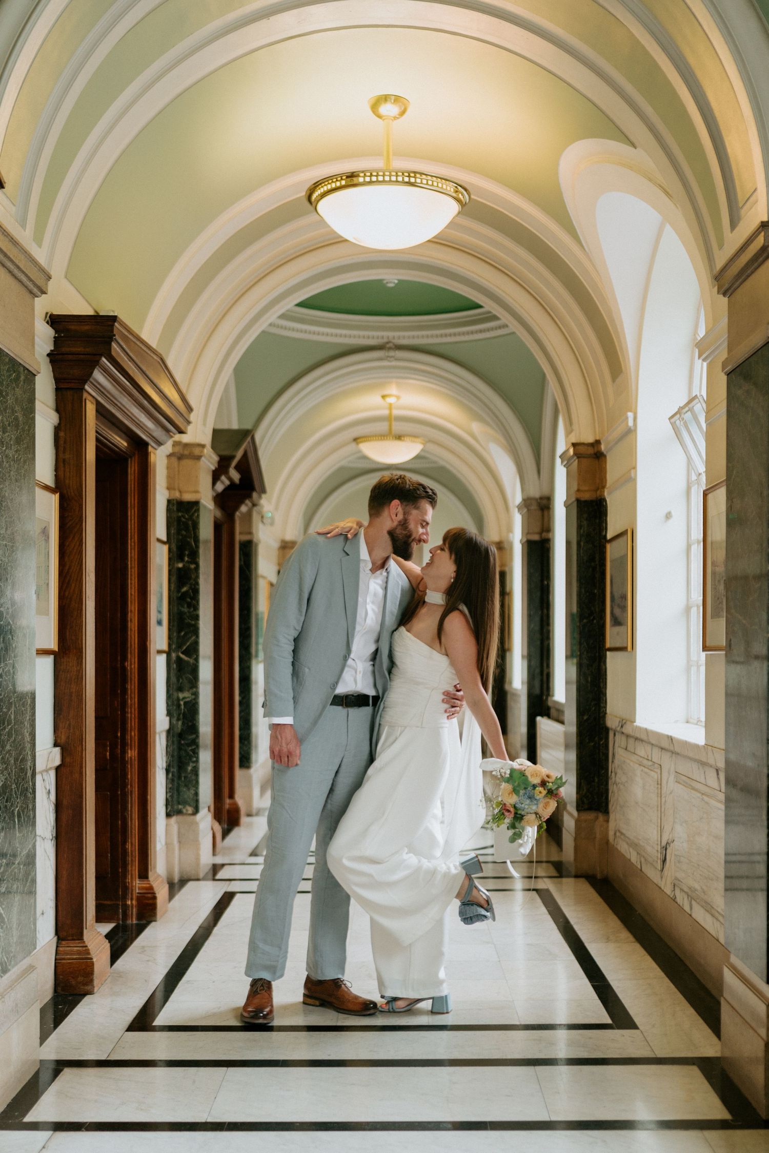 Elegant couple embraces in a historic marble hallway with arched ceilings and ornate architectural details.