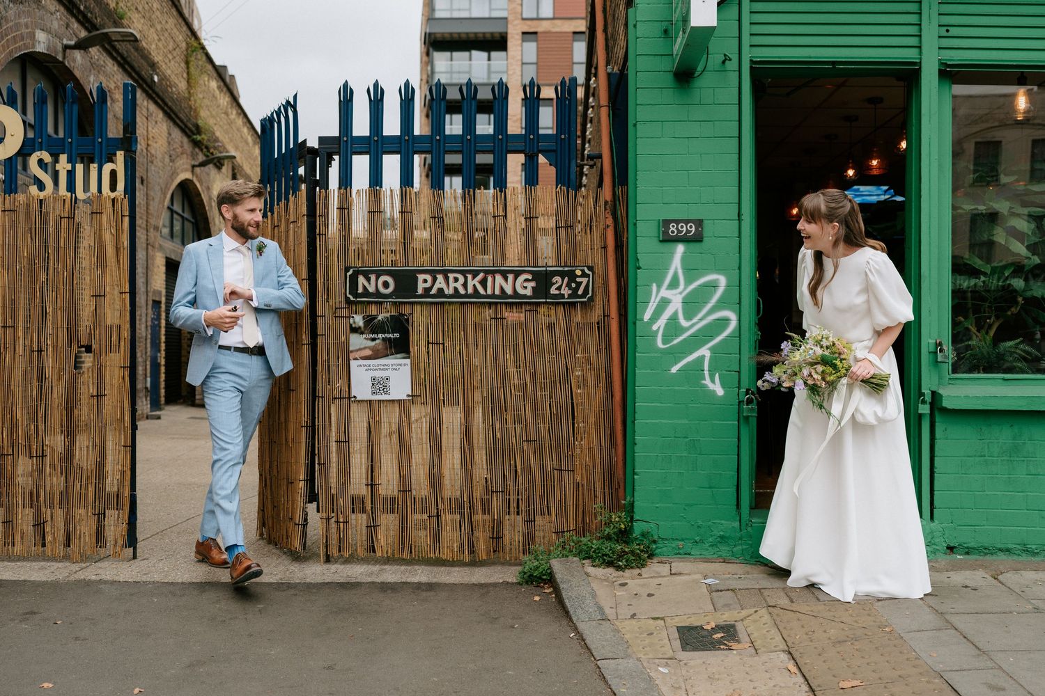 Wedding couple poses in front of rustic wooden walls and green storefronts on an urban street.
