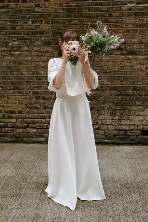 A figure in a flowing white wedding dress holds up a vintage camera while holding a colorful bouquet of flowers against a brick wall.