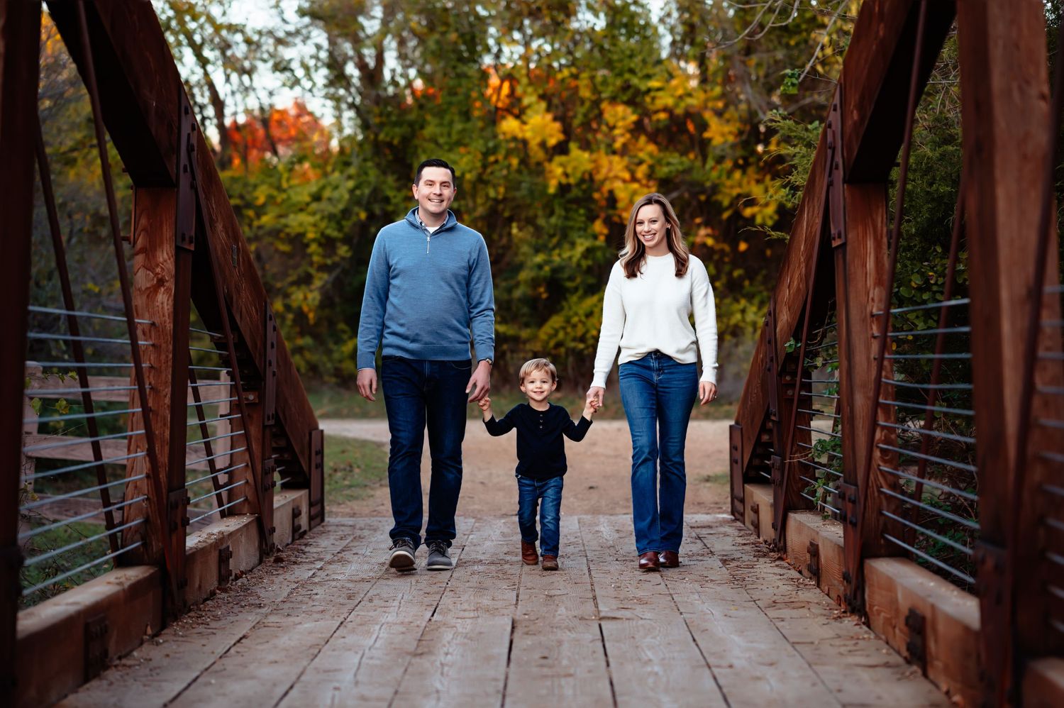 mom and dad hold toddler son's hands while walking across wooden bridge during outdoor fall familiy photo session