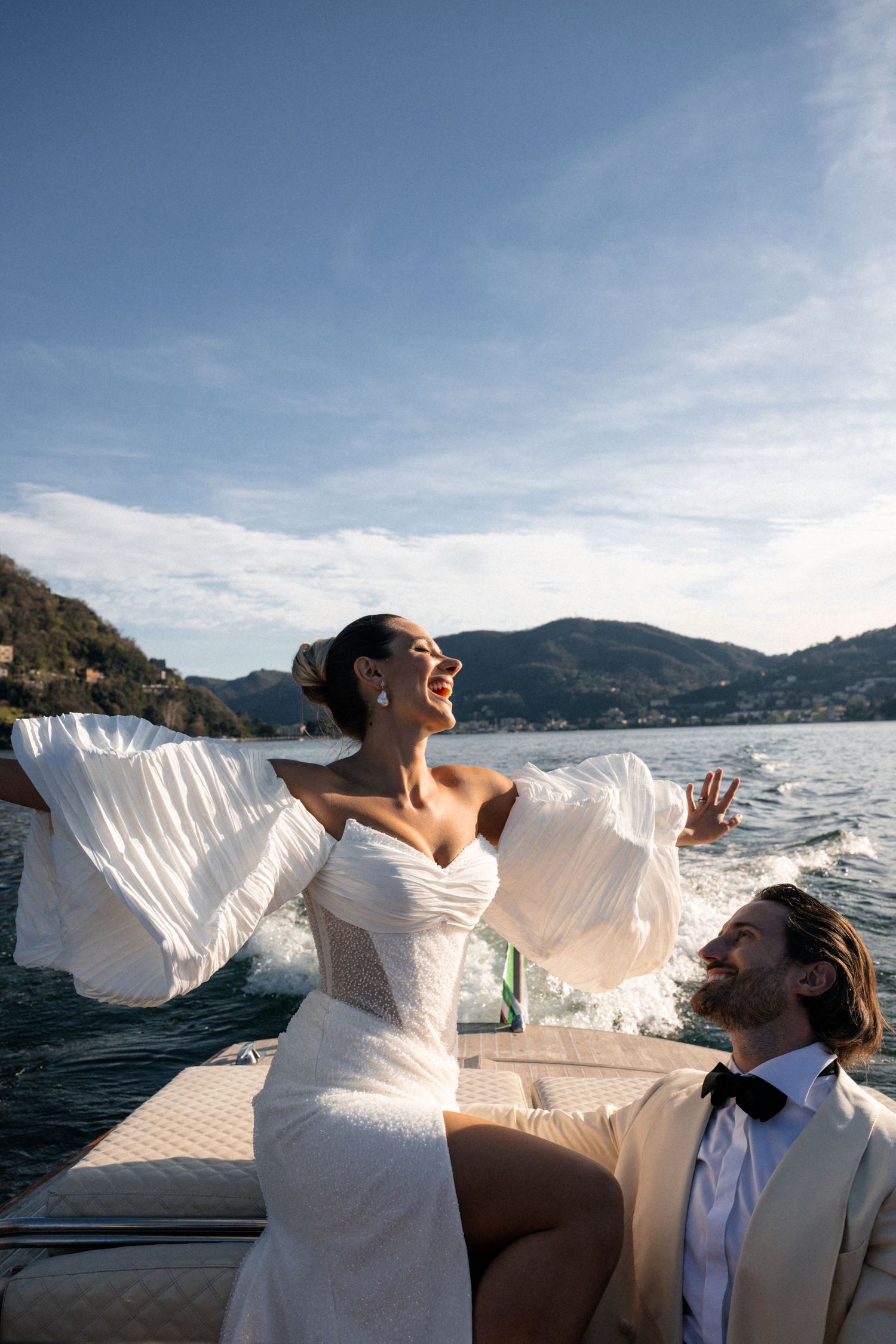 A couple embraces on a pebbly beach with palm trees and hillside buildings in Nice, France.