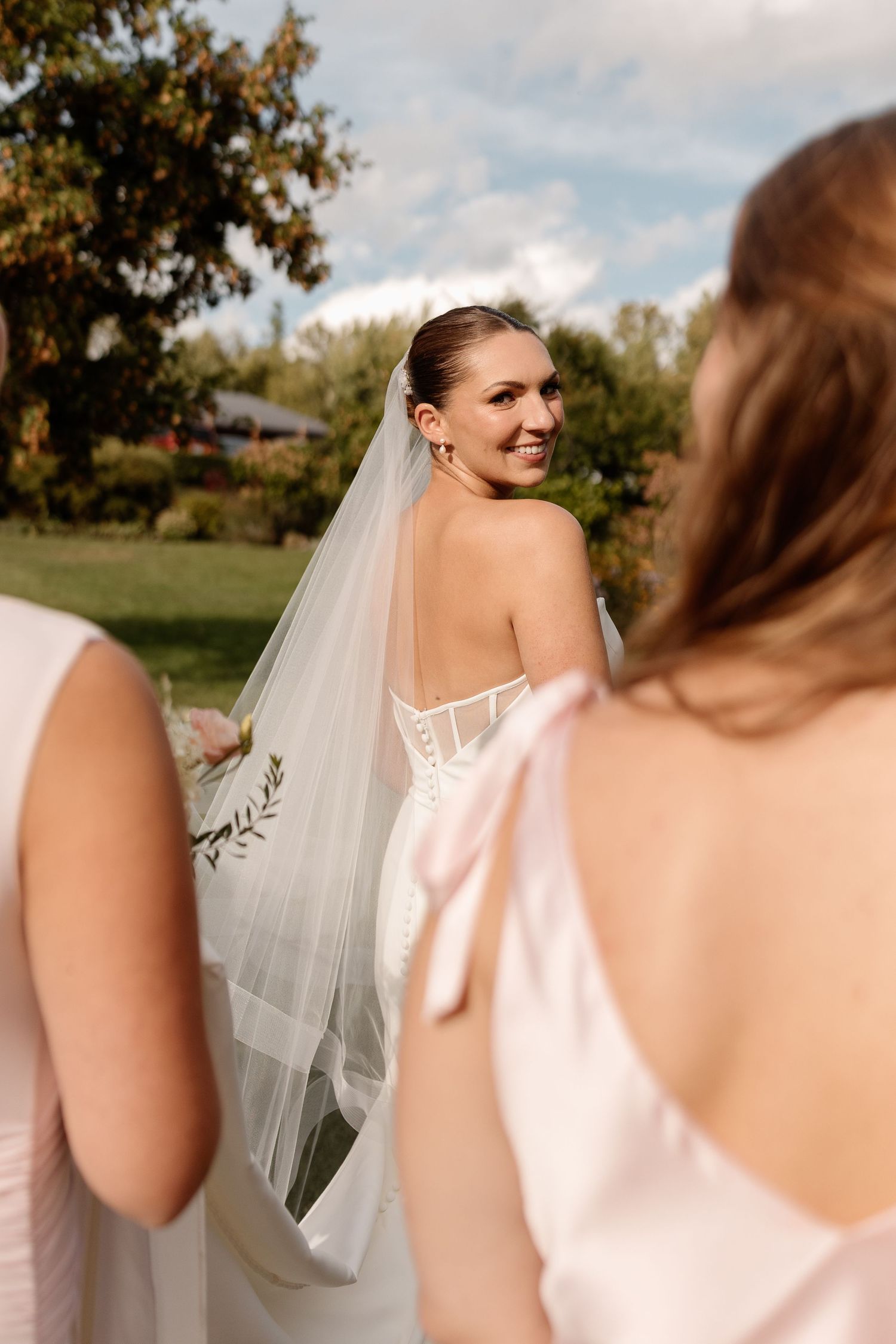 A bride and bridesmaid share a candid moment outdoors on a sunny autumn day, with soft pink dresses and a flowing veil.