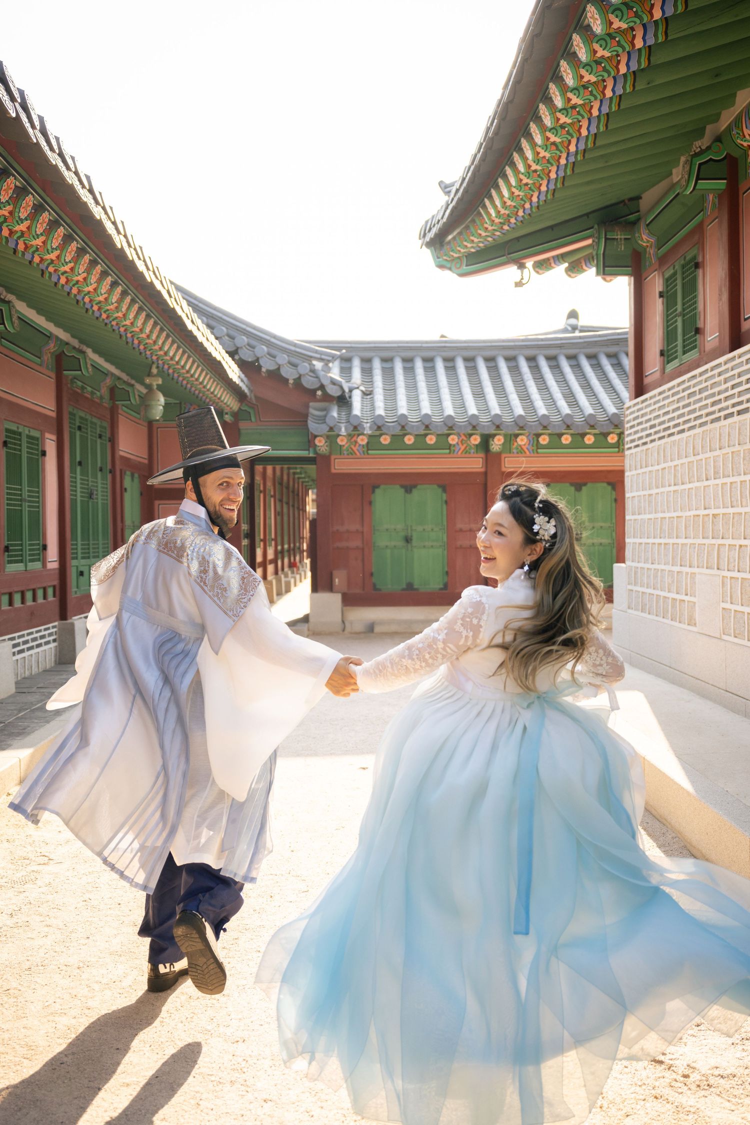 A couple in traditional hanbok clothing dances playfully at a historic Korean palace.