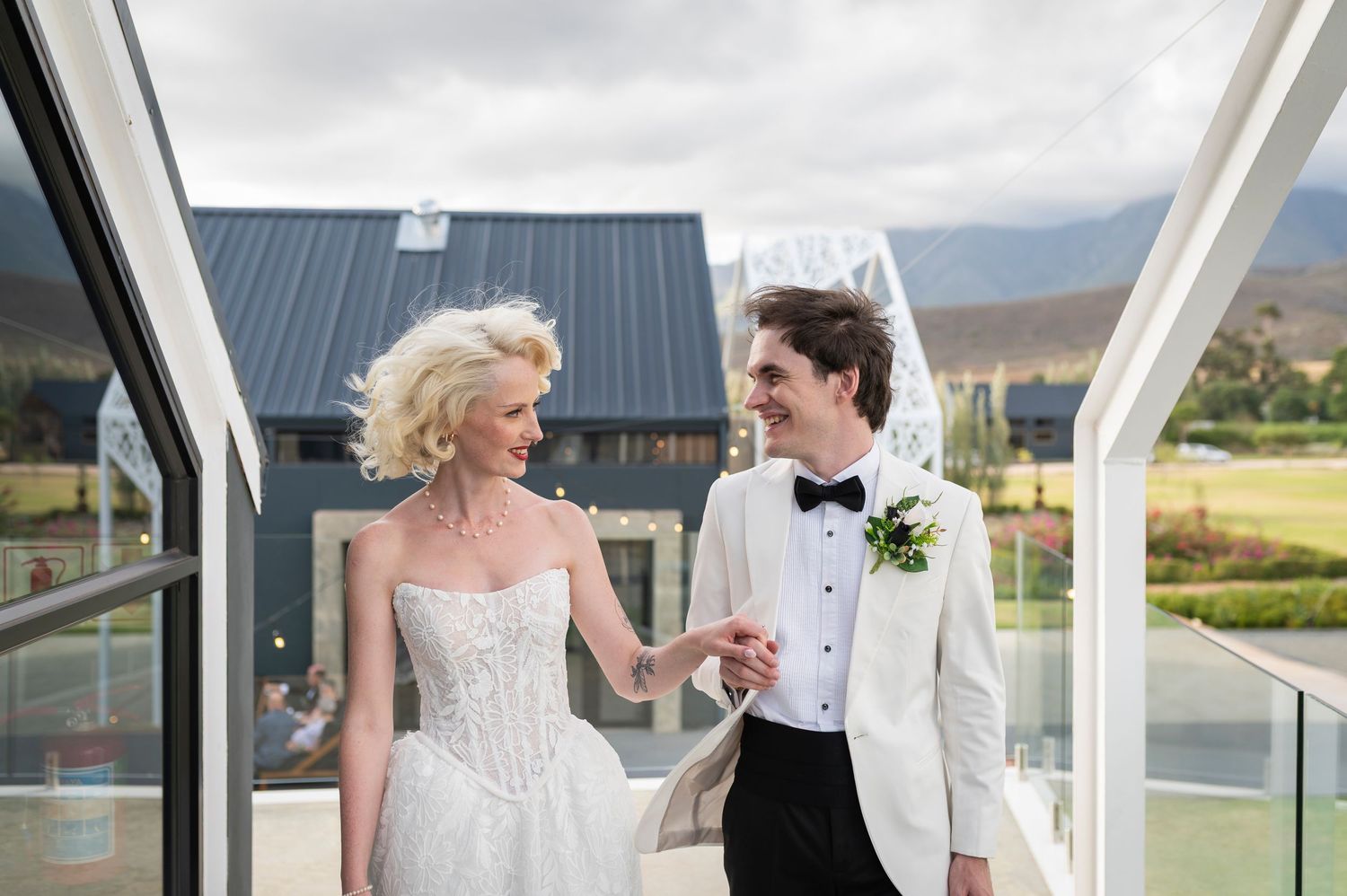 A couple in formal wedding attire share a joyful moment under a covered walkway with mountains in the background.