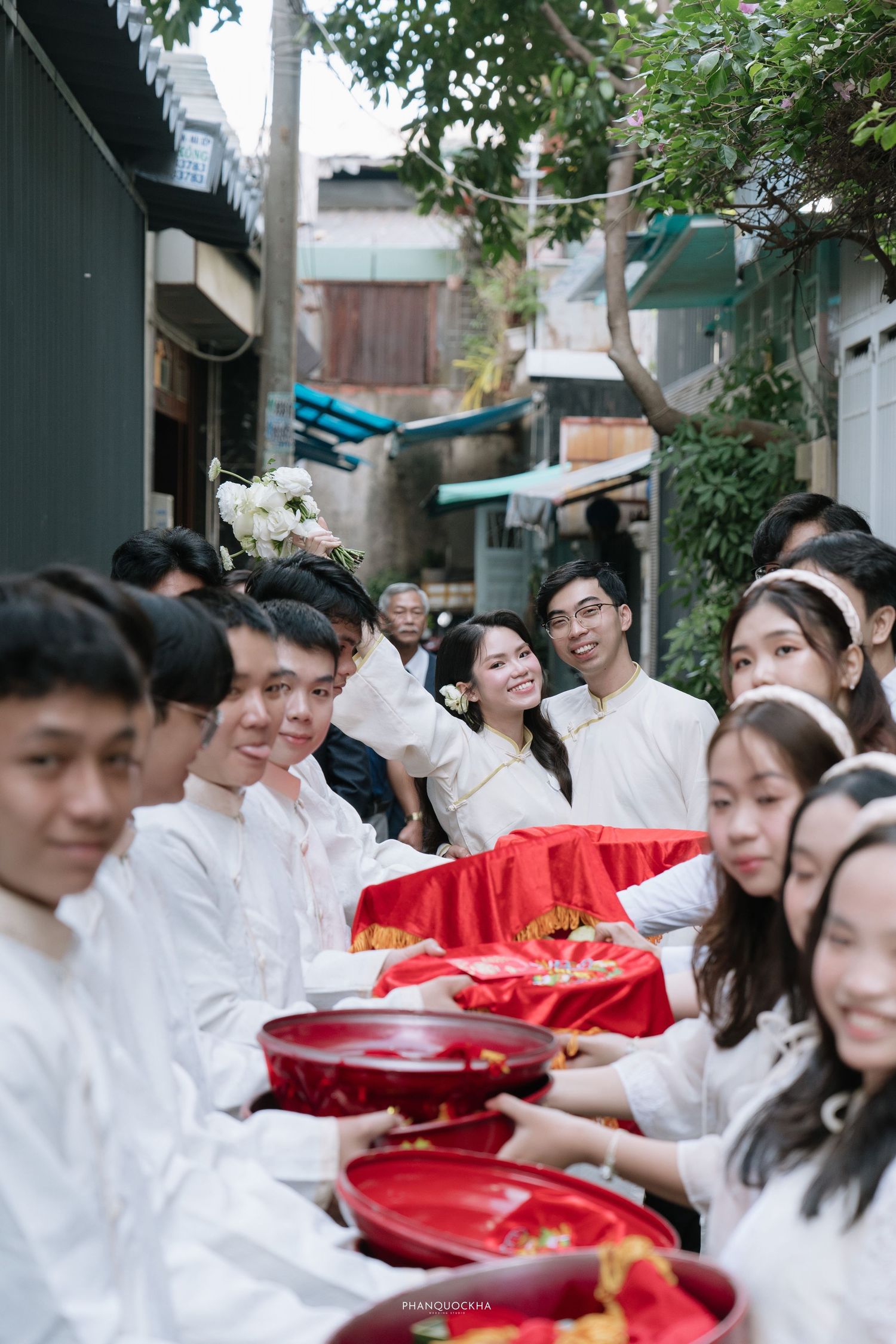 Smiling Vietnamese bride in white ao dai during traditional wedding procession with gift bearers.