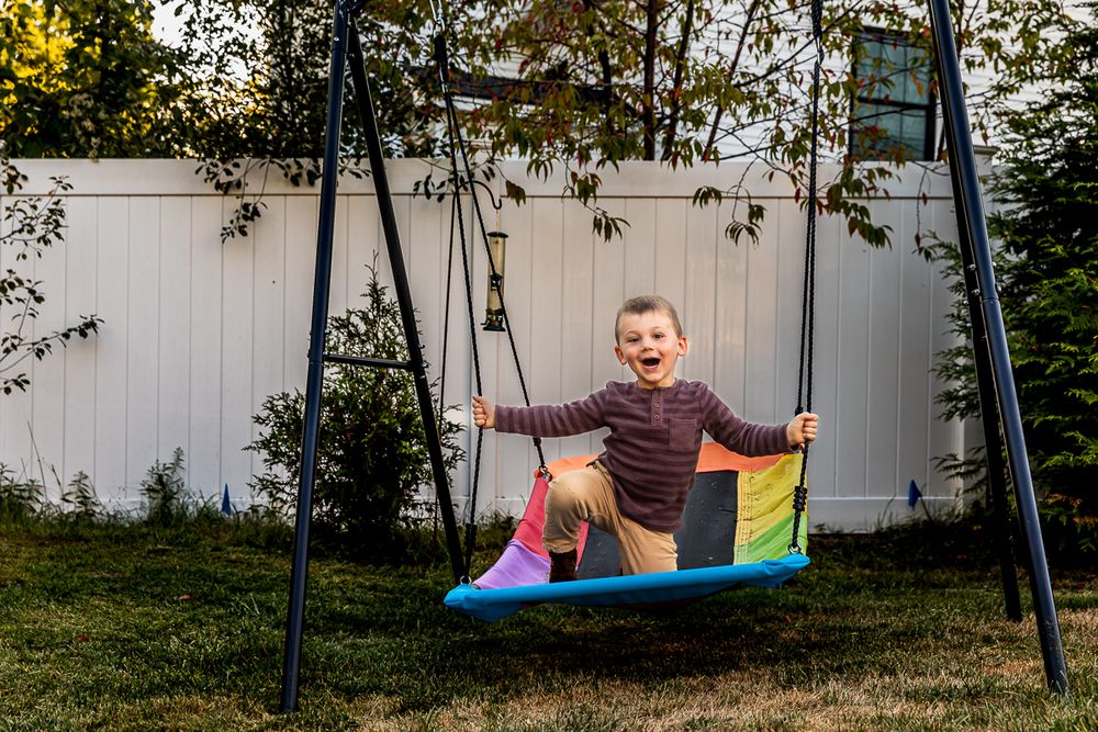 A young child enjoys swinging on a colorful rainbow swing in a backyard setting.