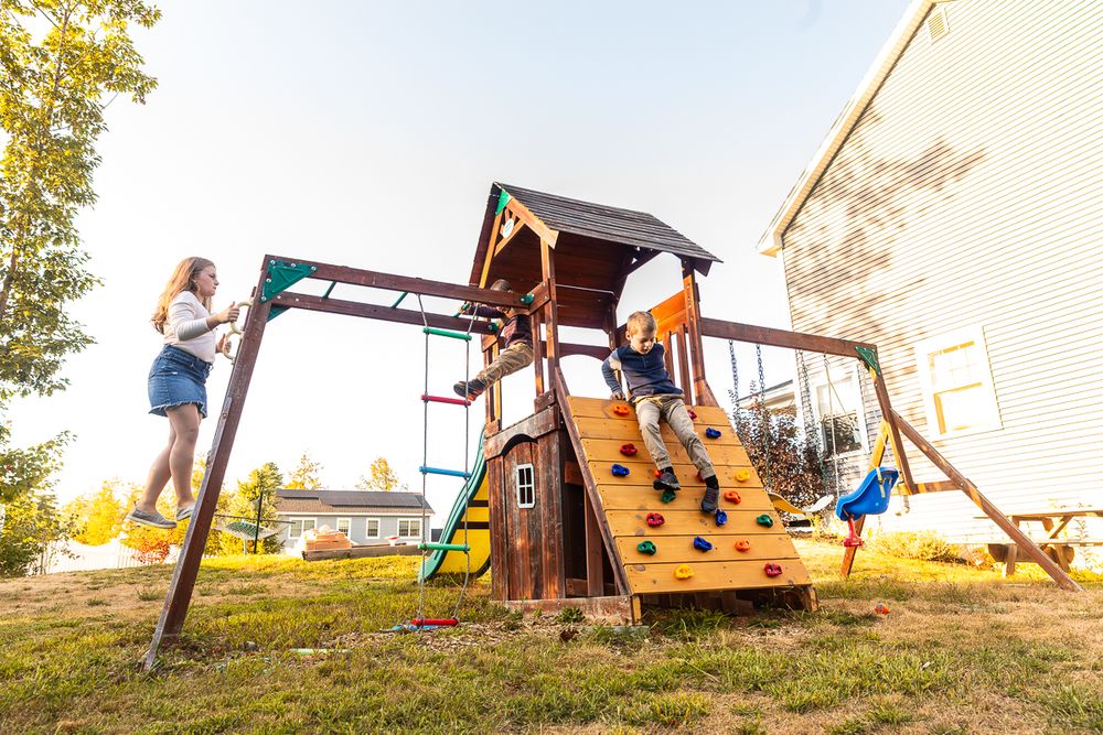 Children play on a wooden backyard playset with swings and slides during a sunny afternoon.