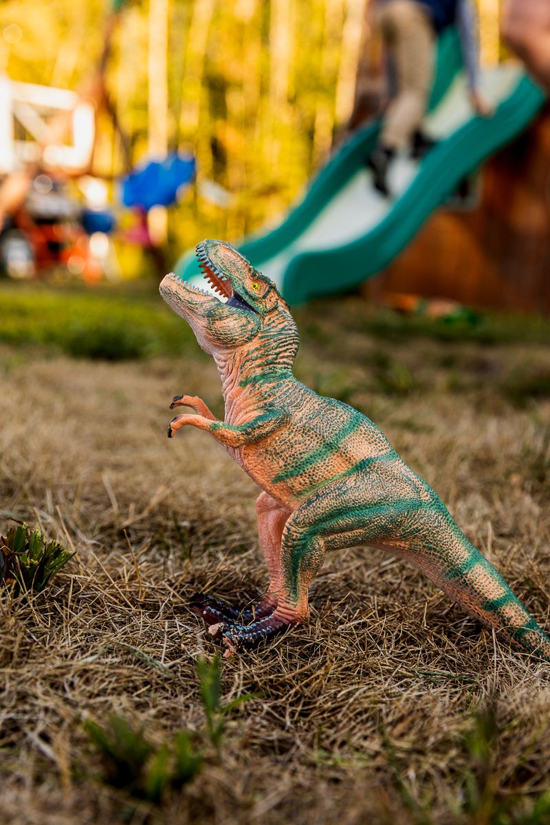 A green plastic toy dinosaur stands in dynamic poses against autumn grass and fallen leaves.