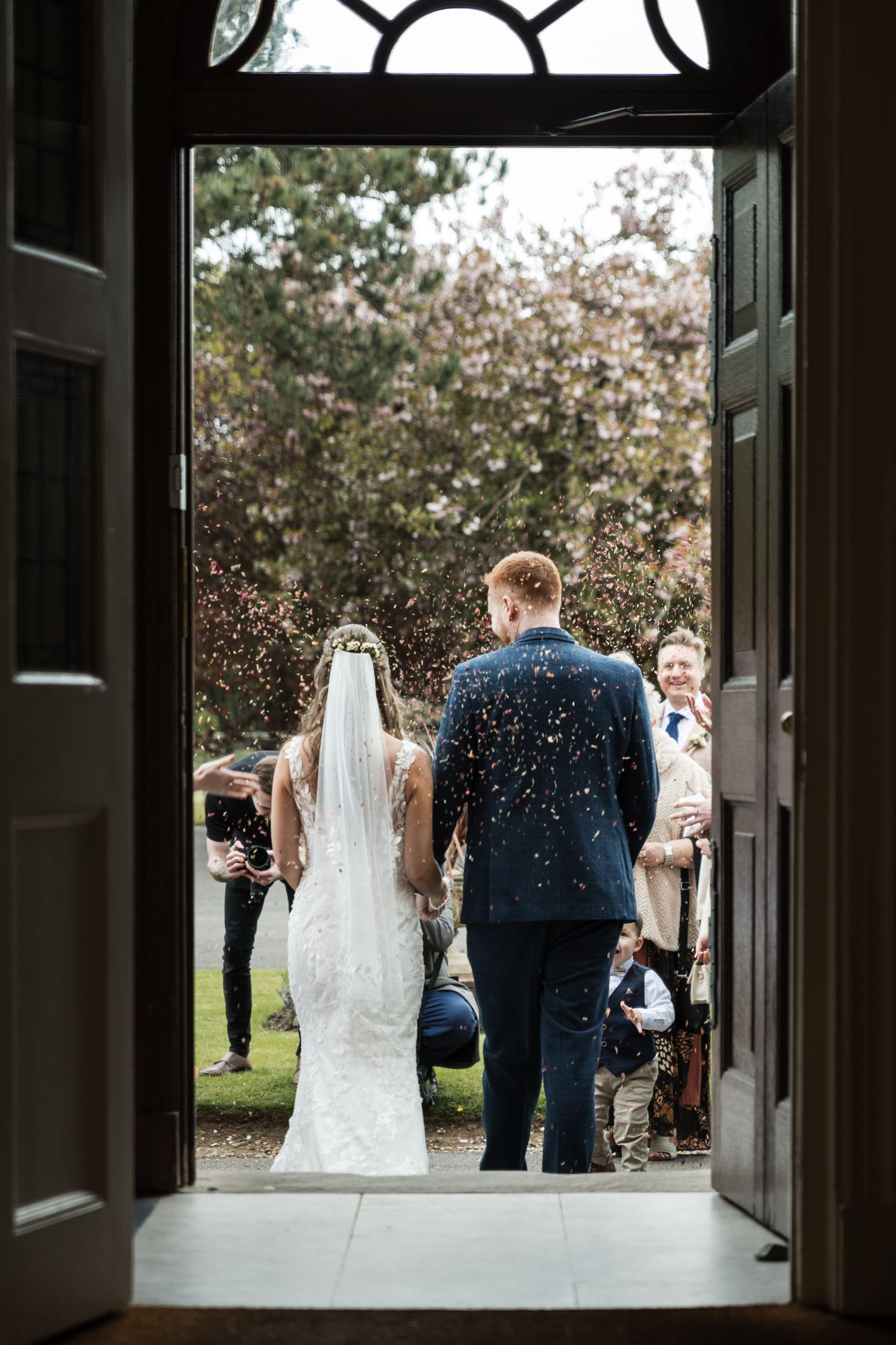 Church doors frame a couple and their wedding party exiting after a ceremony as flower petals cascade down.