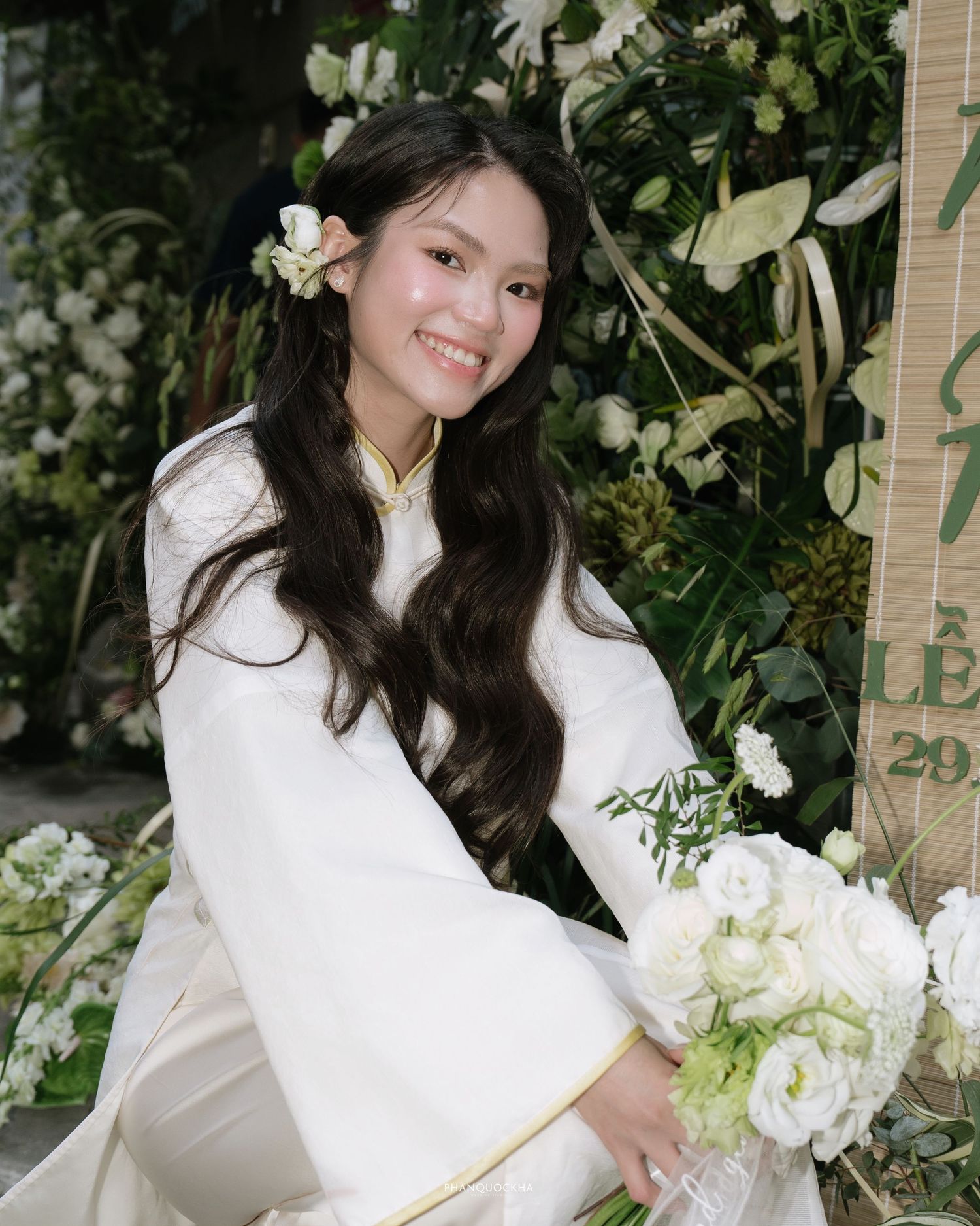Beautiful Vietnamese bride in white ao dai smiles holding white bouquet beside floral wedding backdrop.