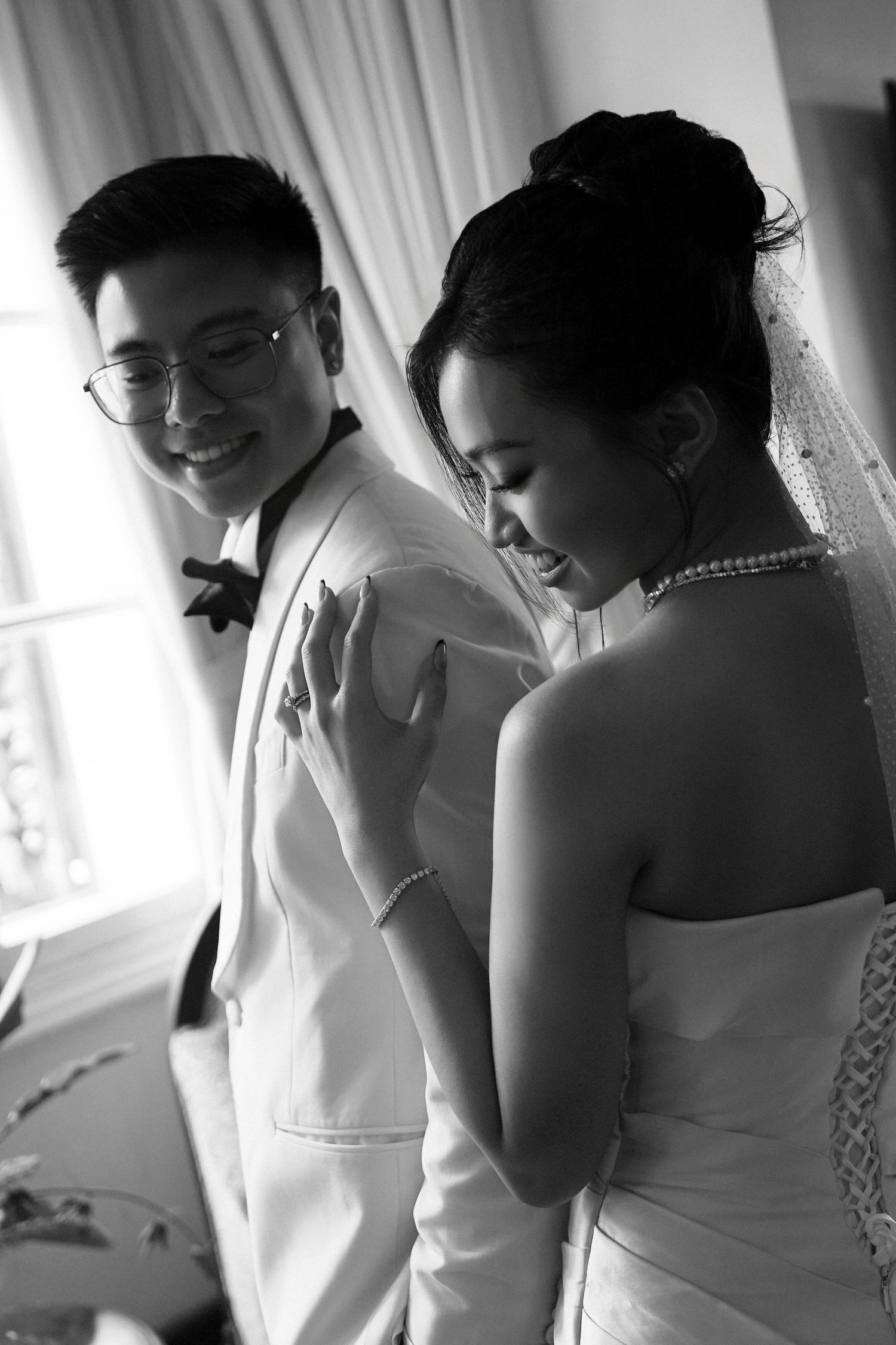 Black and white photo of smiling Asian groom in white suit with bride in strapless wedding dress indoors.