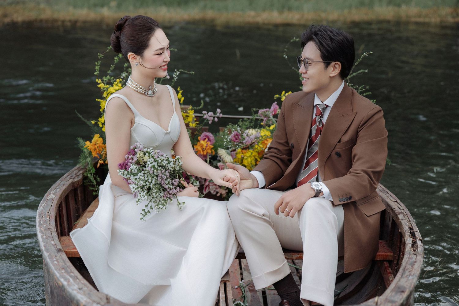 Bride in white gown and groom in brown suit holding hands in a floral boat on a tranquil river.