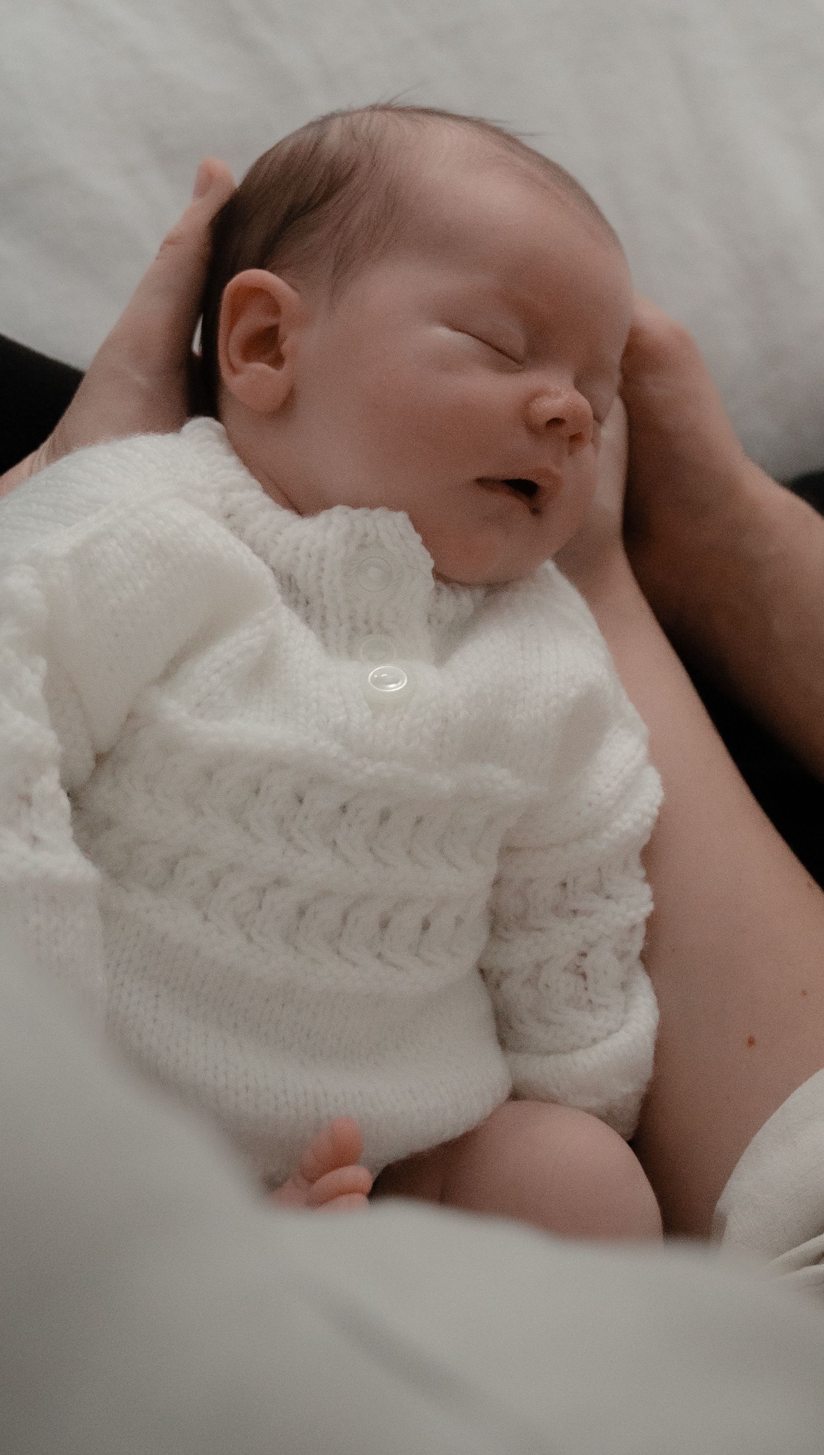 A sleeping newborn baby wearing a soft white knitted sweater rests peacefully in gentle arms.