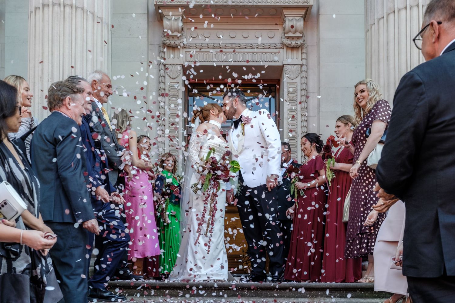 Wedding guests throw confetti at a couple outside an elegant columned building entrance during a joyous celebration.
