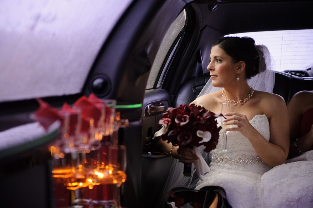 A bride wearing white sits in a limousine with candlelight and a burgundy bouquet.