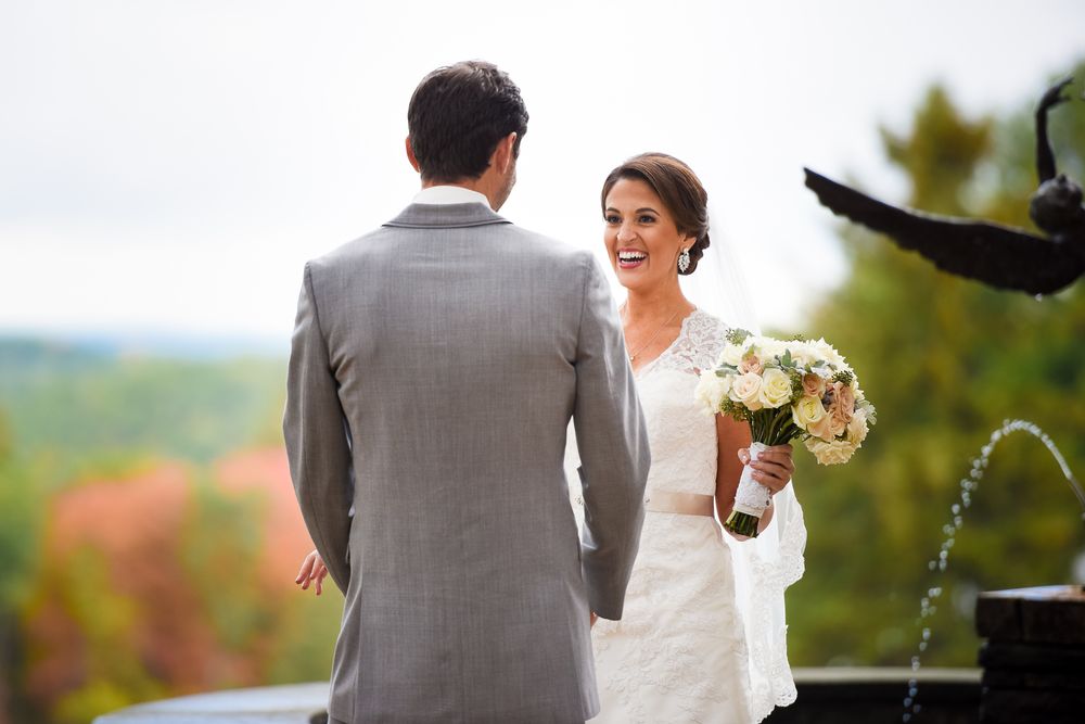 Wedding couple stands together outdoors with autumn foliage and mountains in background as bride holds white bouquet.