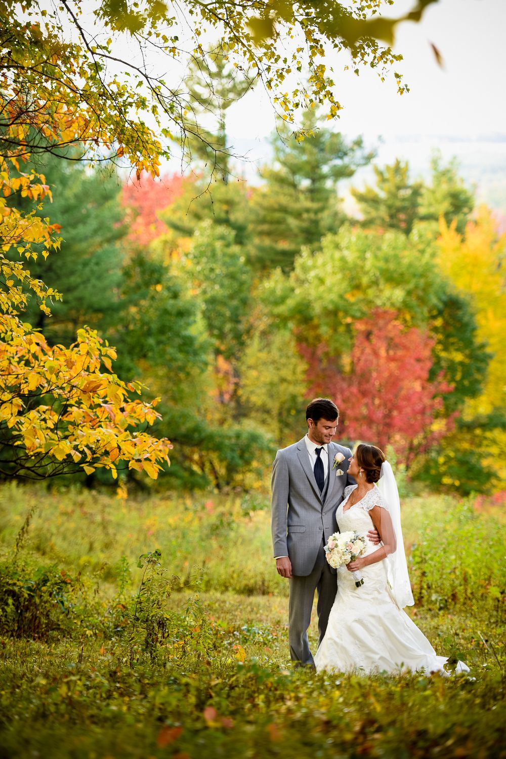 Wedding couple walks through vibrant autumn foliage with yellow and red trees in sunlit garden setting.
