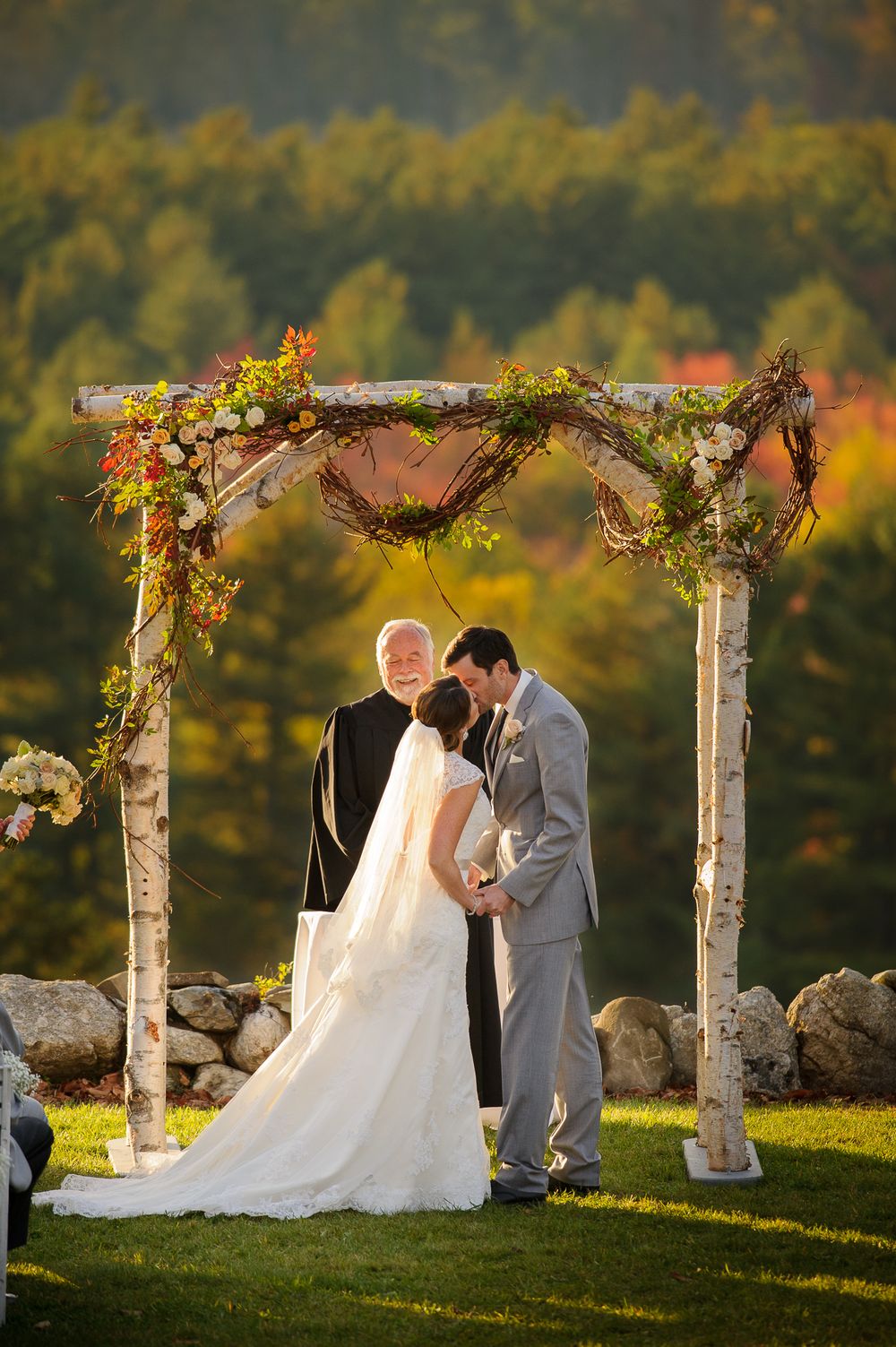 A romantic autumn wedding ceremony takes place under a birch archway decorated with flowers and foliage at sunset.