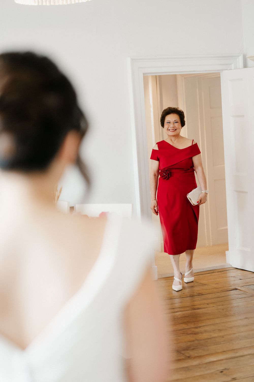 Woman in elegant red off-shoulder dress poses indoors against white walls and wooden flooring.