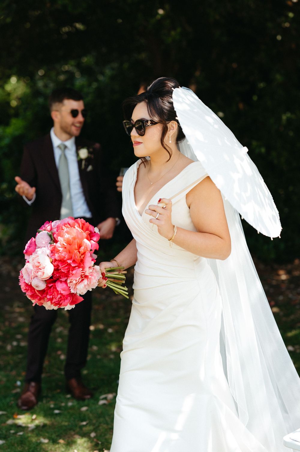 Wedding portrait outdoors with white bridal gown, parasol and vibrant pink bouquet in garden setting.