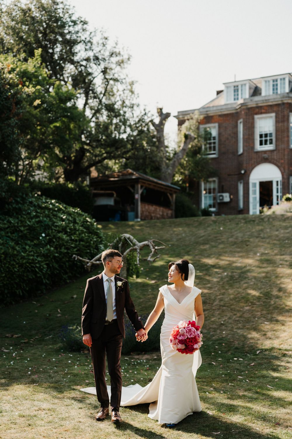 Outdoor wedding couple walking and smiling together on a grassy hillside with brick houses in the background.