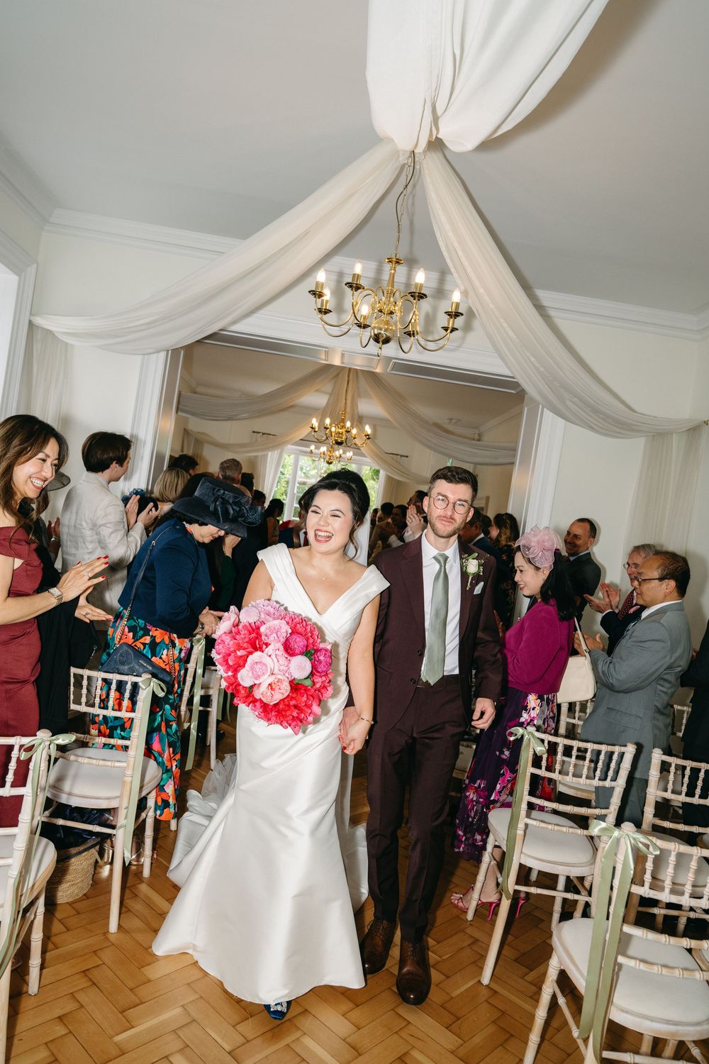Wedding guests dance and celebrate under elegant white draped fabric and chandeliers in a reception hall.