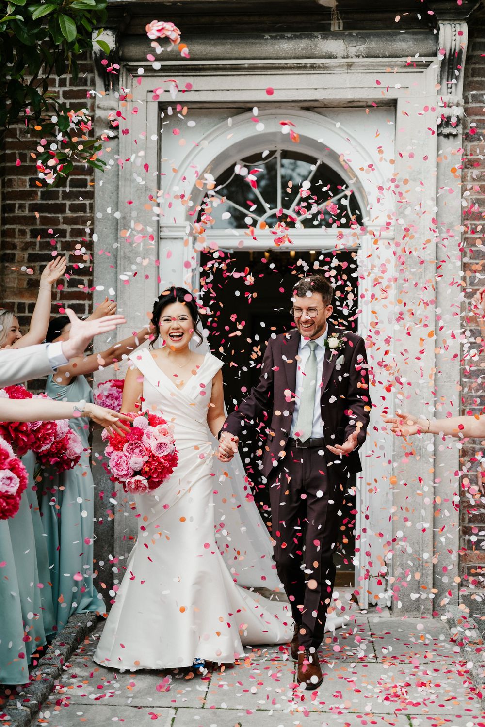 Wedding couple celebrates with pink confetti being showered over them outside a brick building entrance.