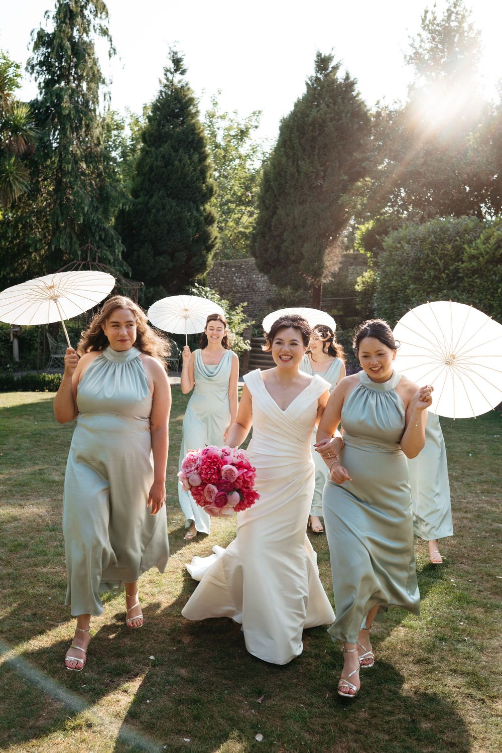 Wedding party holding ornate white parasols poses playfully in sunlit garden during summer celebration.