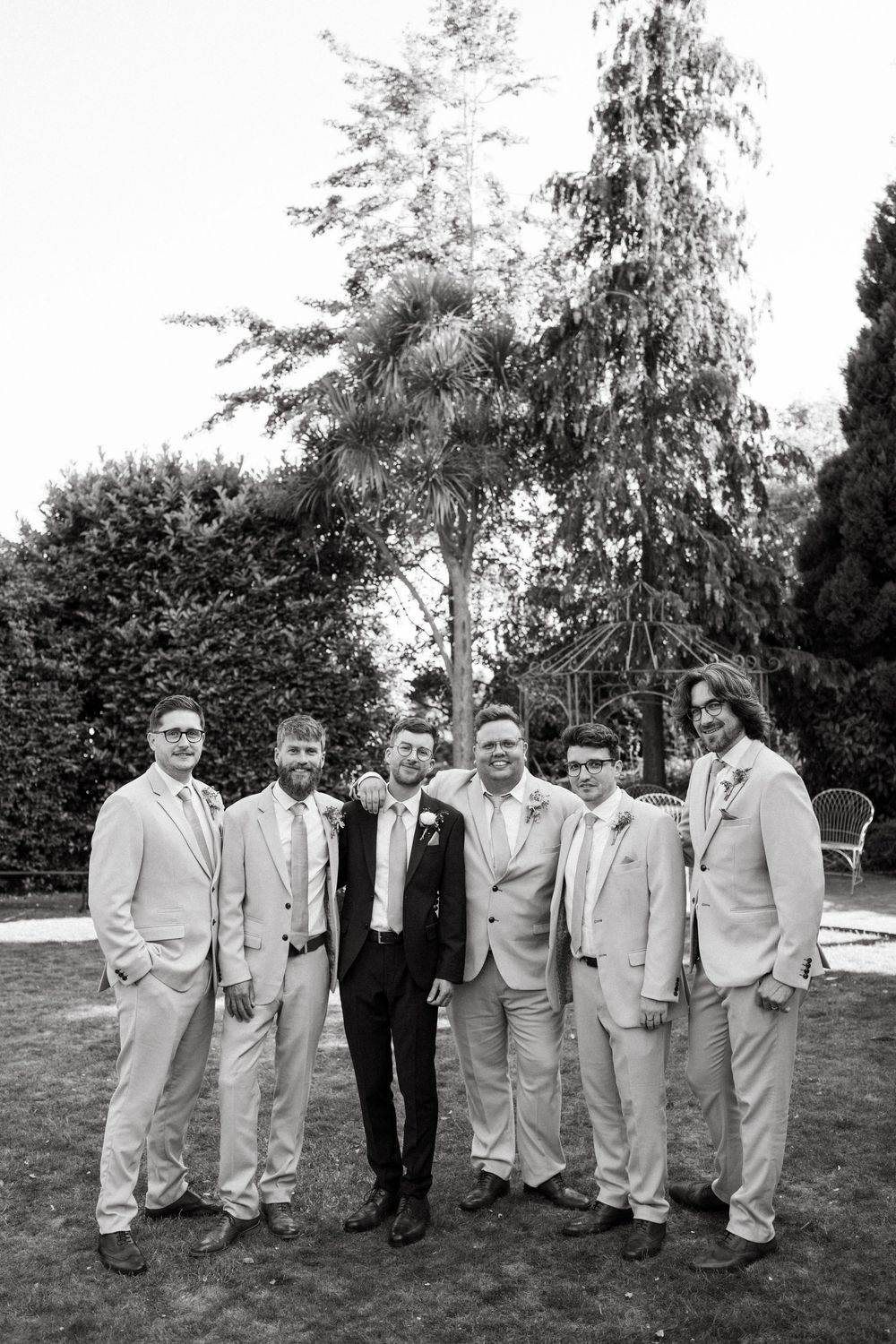 Wedding party members in light gray suits and black attire pose together on a lawn with evergreen trees.
