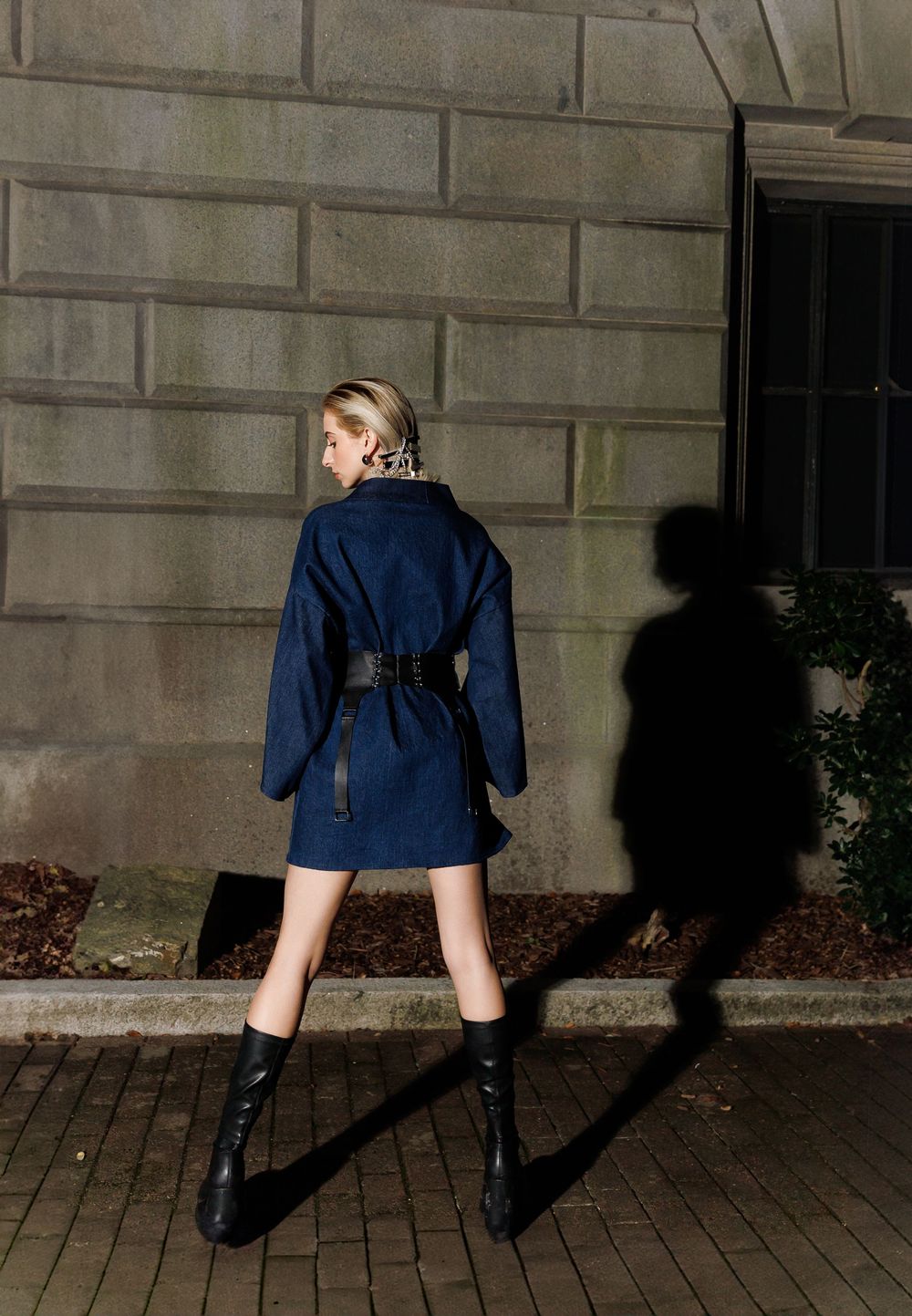Fashion model wearing navy blue coat dress and tall boots poses dramatically against stone wall casting long shadow.