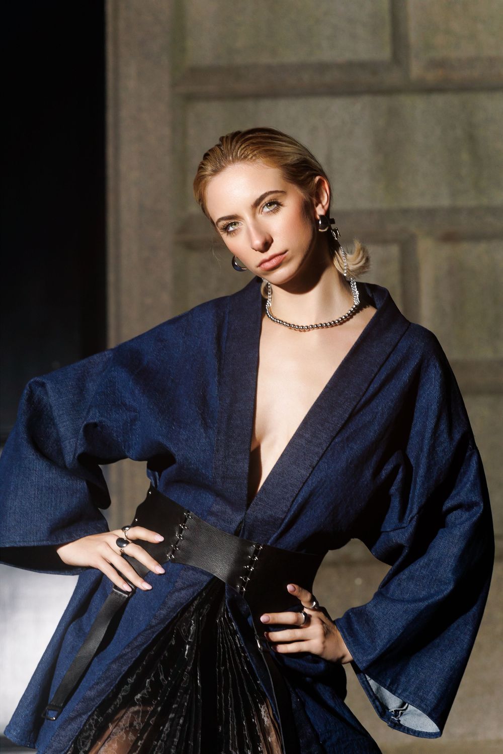 Model in navy blue jacket with plunging neckline and black corset belt poses against textured wall in studio lighting.