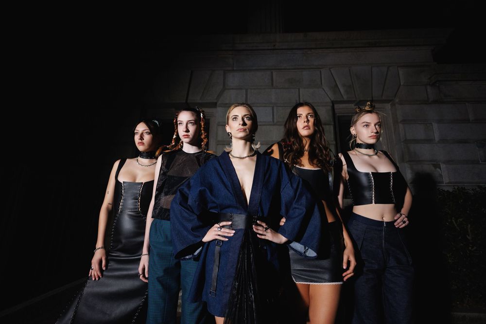 Group wearing casual and formal outfits poses together against dark textured wall in dramatic lighting.