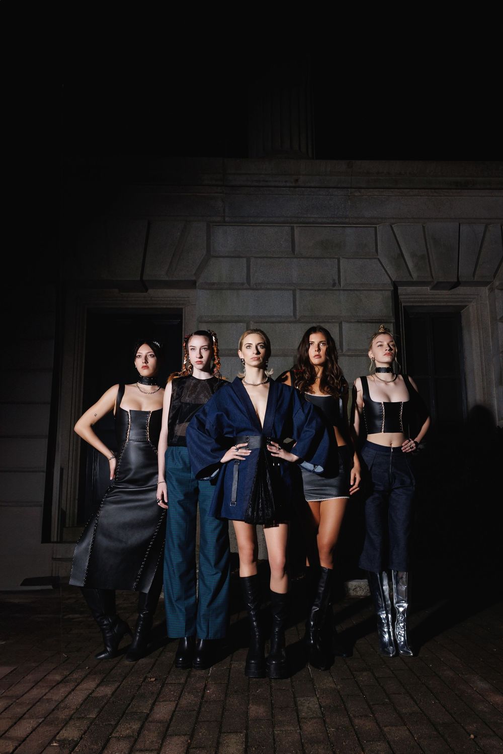 Group in dark formal attire poses against brick wall in dimly lit industrial space.