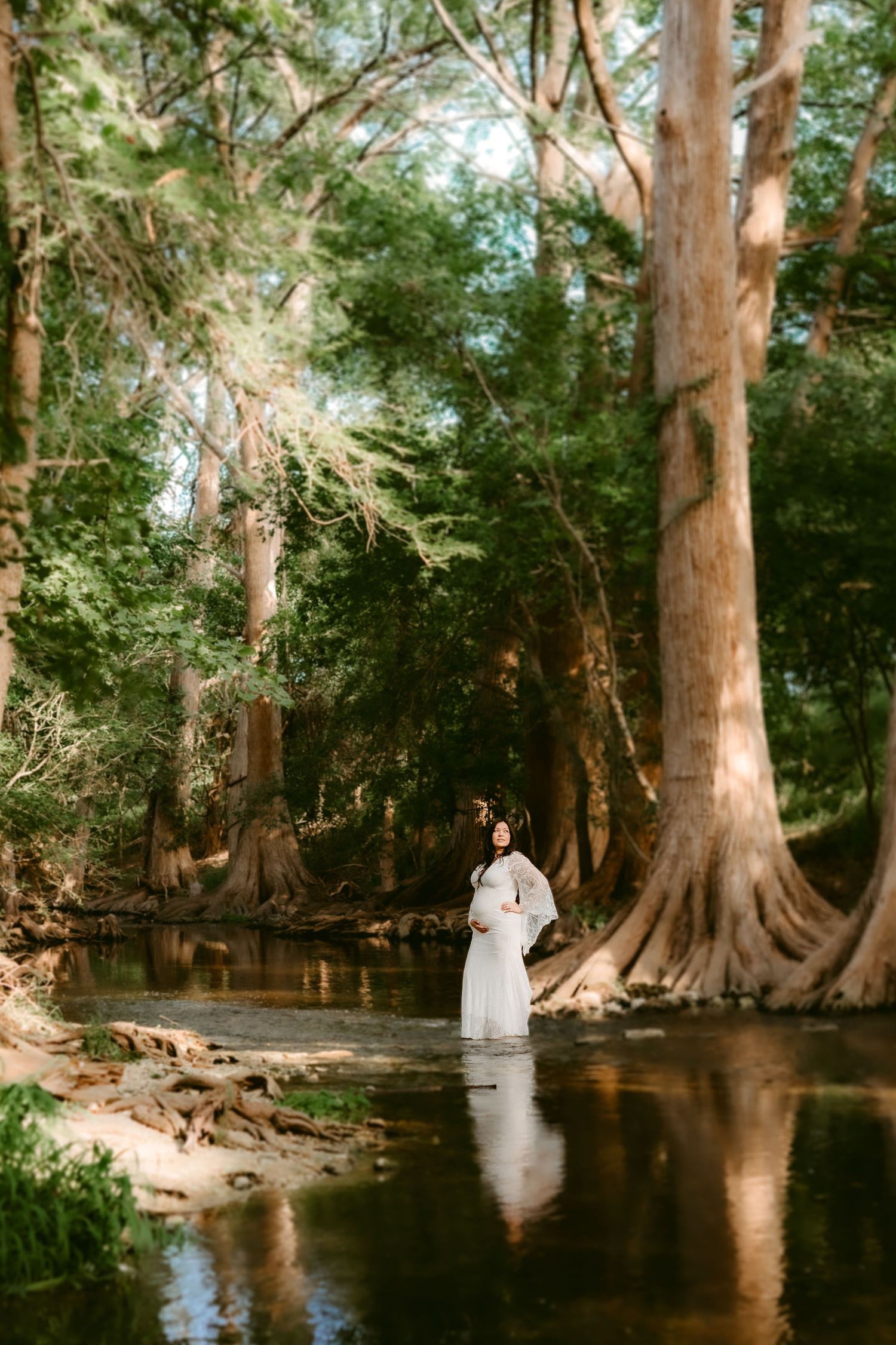 Person in white dress stands in tranquil cypress-lined river with sunlight filtering through trees in ethereal forest scene.