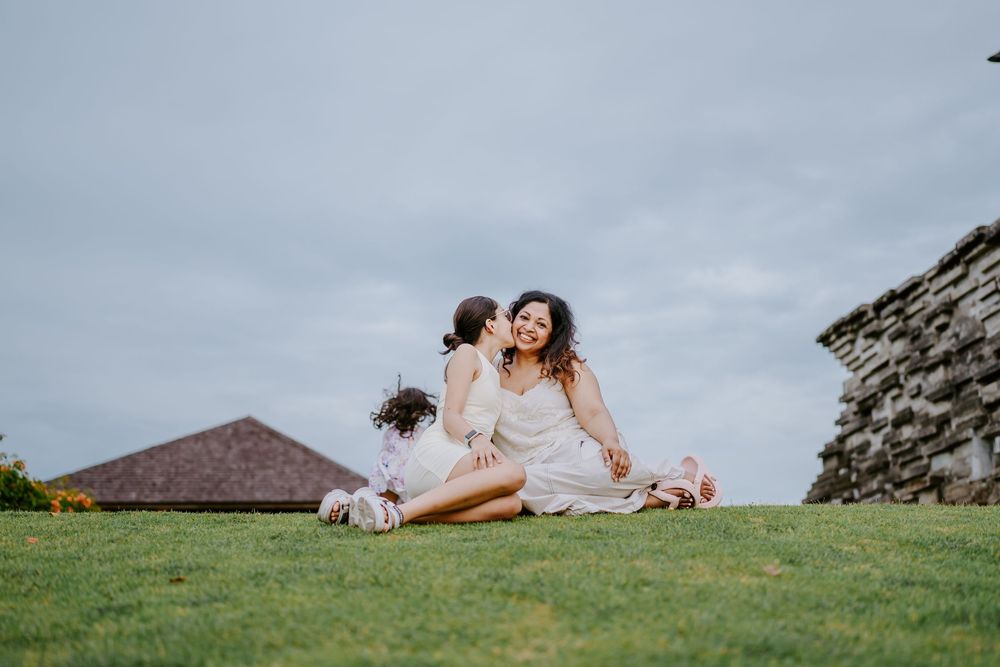 Two people in white dresses share joyful moment while sitting on grass under cloudy sky.