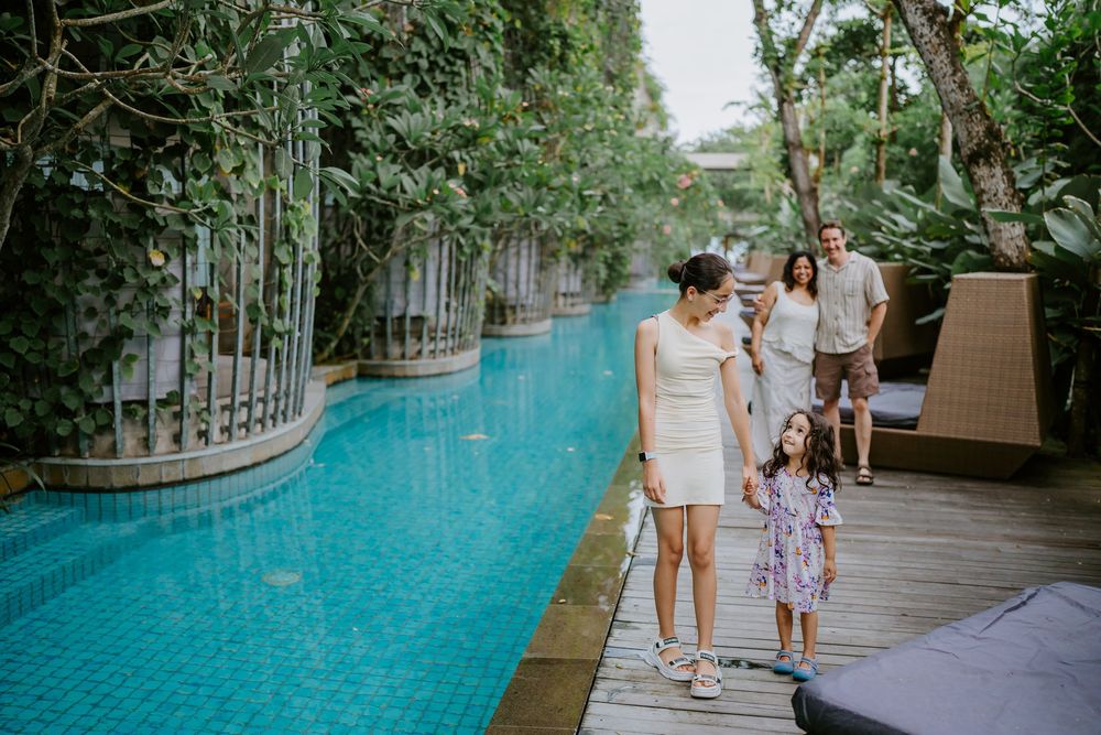 A family strolls along a wooden walkway beside a turquoise swimming pool.