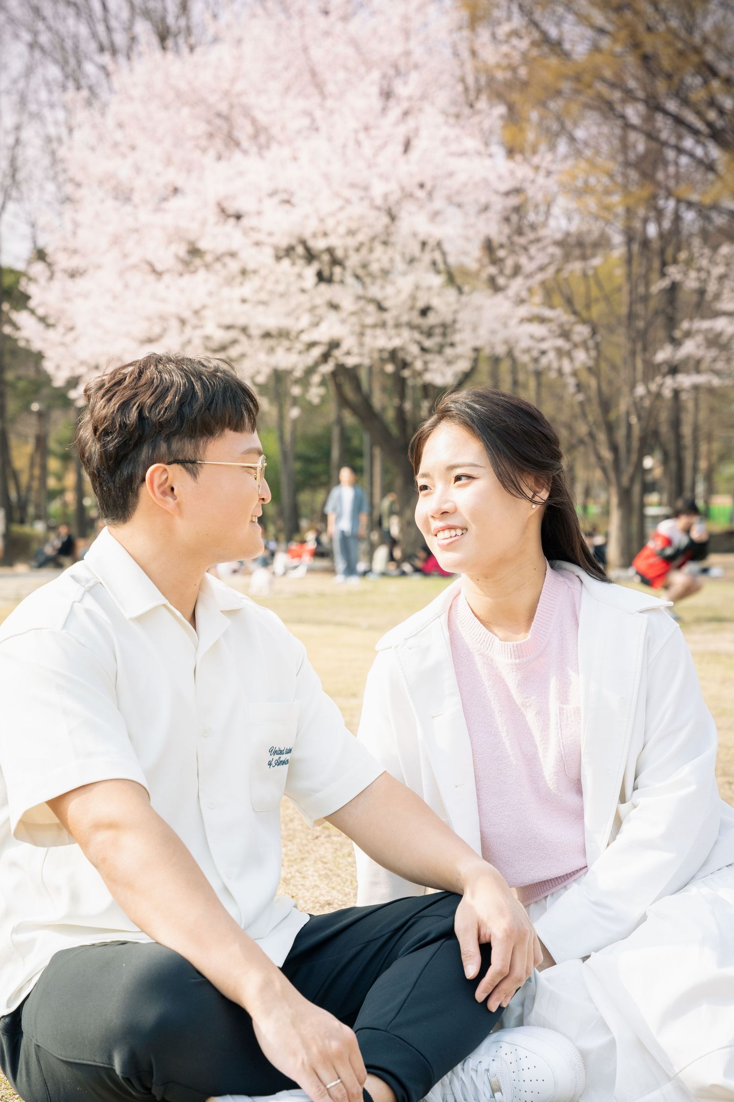 A couple sits together on a sunny spring day with blooming cherry blossoms in the background at a park.