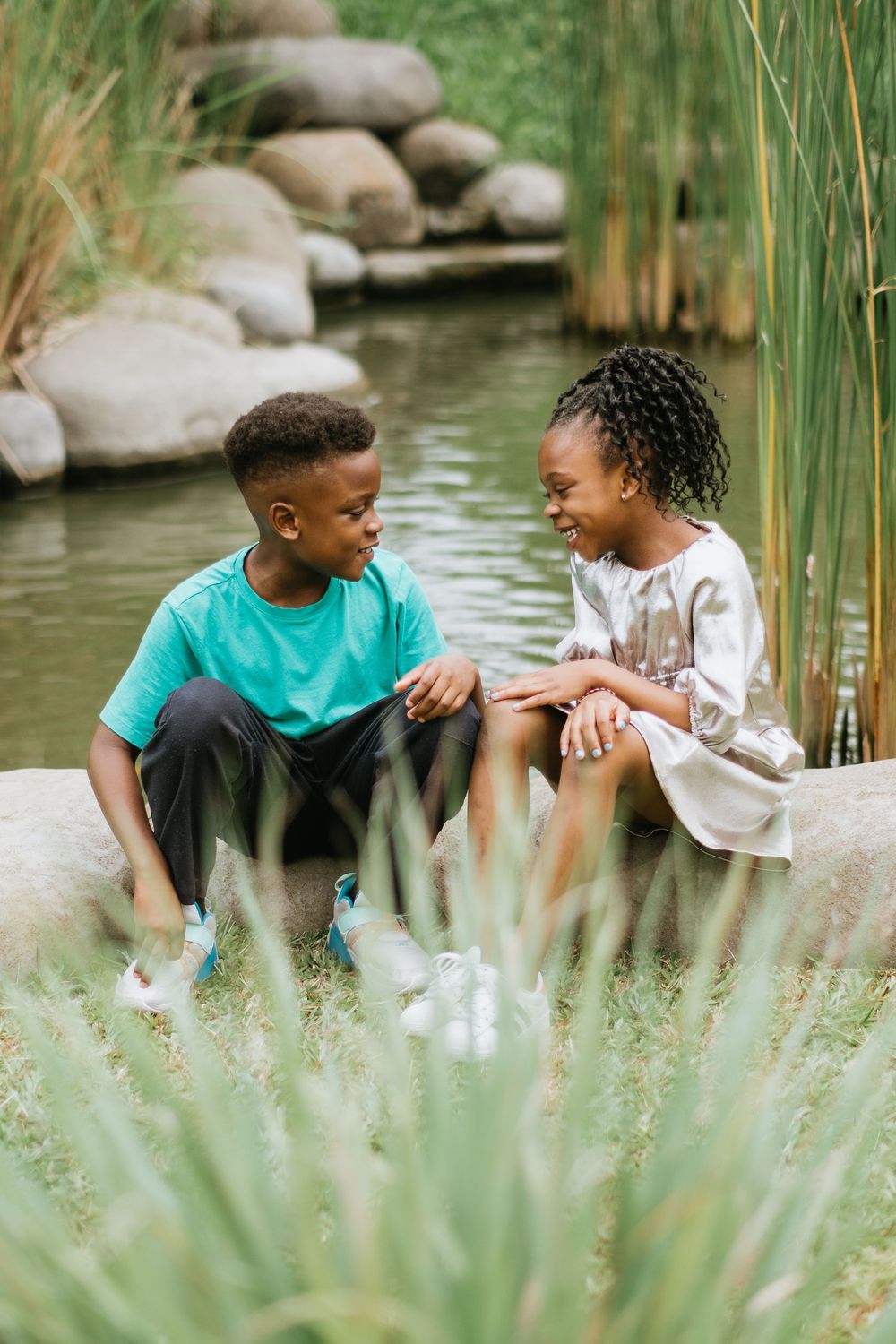 Two children share a sweet moment while sitting by a pond with rocks and reeds.