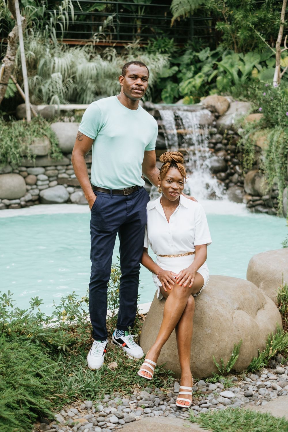 A couple poses by a waterfall and turquoise pool in a tropical garden setting.