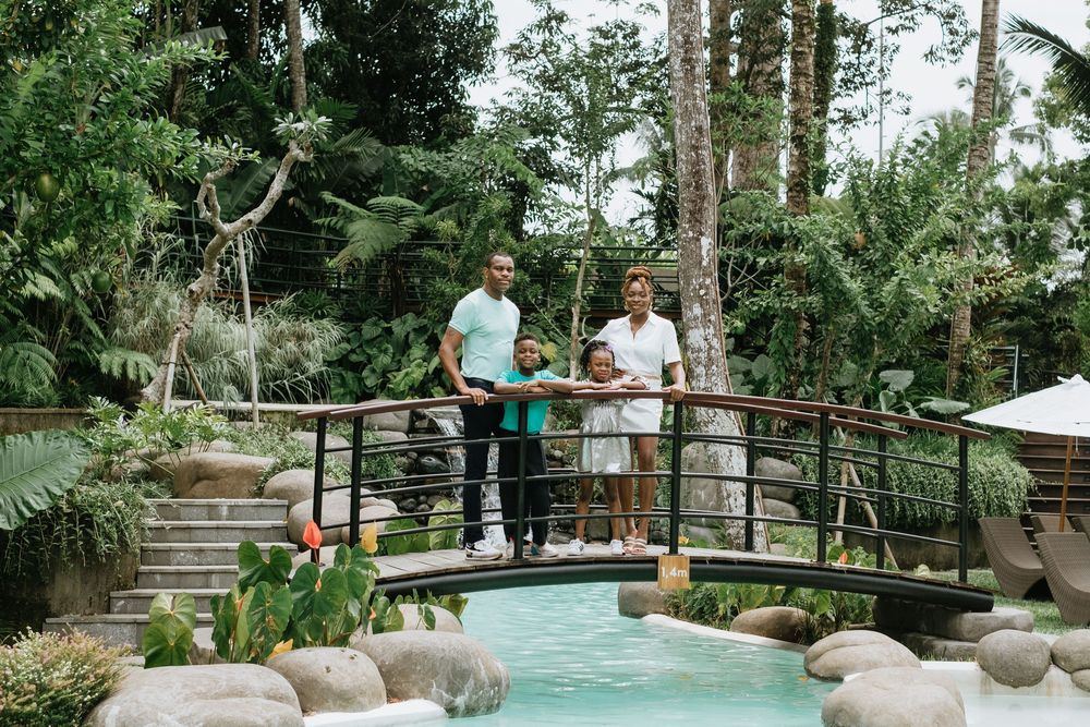 Two people stand on a wooden bridge overlooking a tropical pool with rocks.
