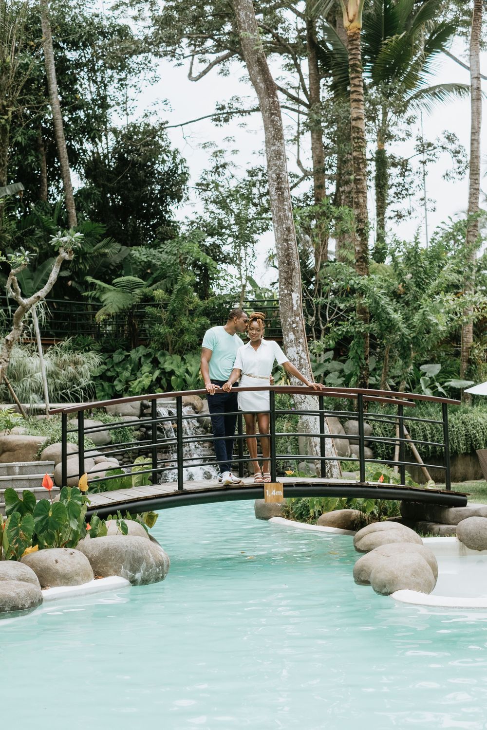 A couple stands together on an arched bridge over turquoise water in a tropical garden.