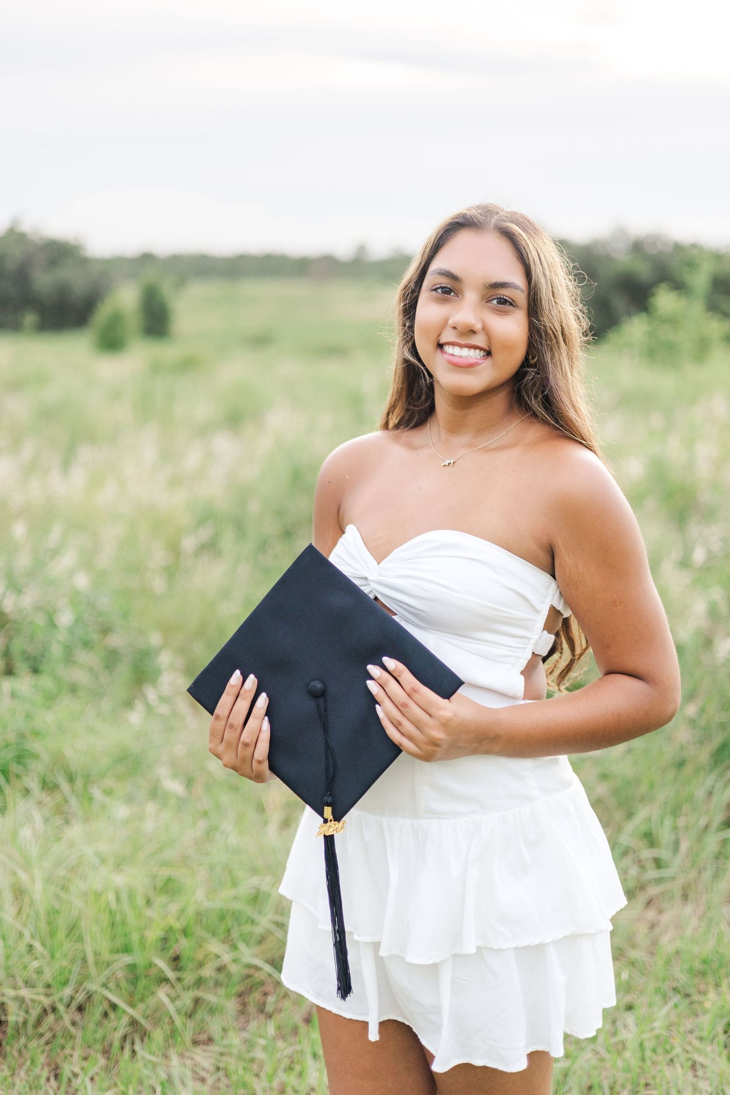 Graduate holding diploma and wearing white dress poses in sunny meadow field.